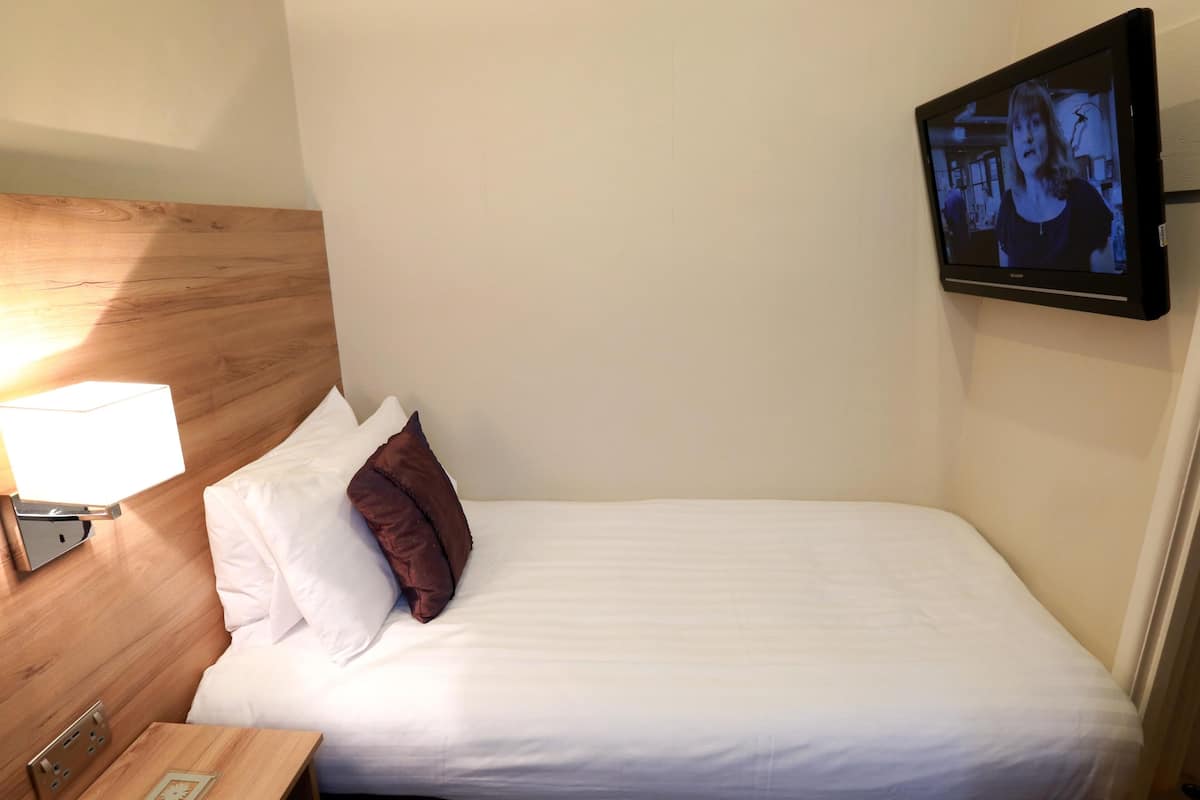 Quarto de hotel com cama, mesa de cabeceira, luminária, TV fixada na parede e tomada.