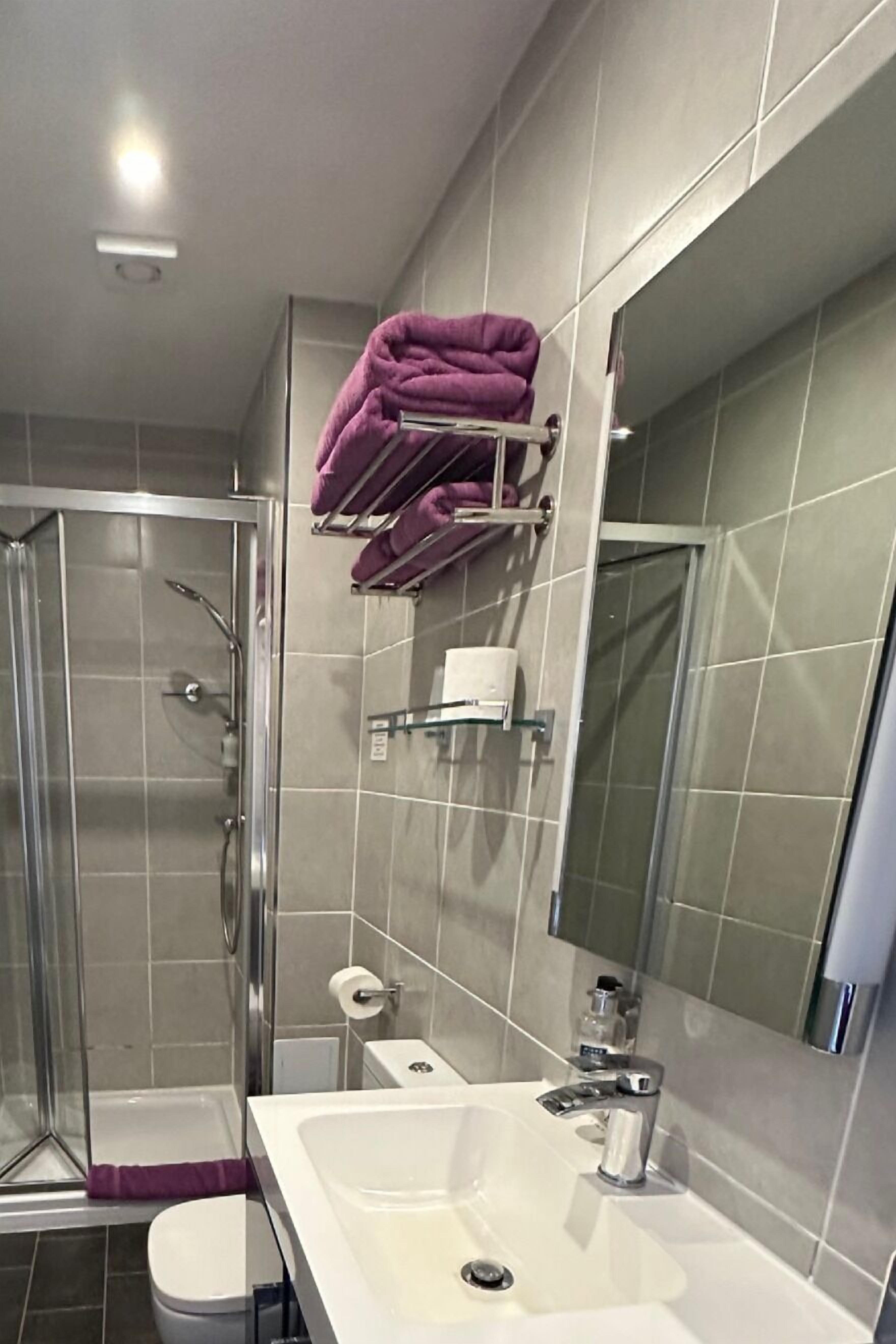 A modern bathroom with a glass-enclosed shower, a white sink with a mirror, and a towel rack with purple towels.