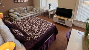 A bedroom with a bed, a sofa, a dining table, a TV, and a window with blinds. - The Chocolate Box Hotel (Bournemouth)