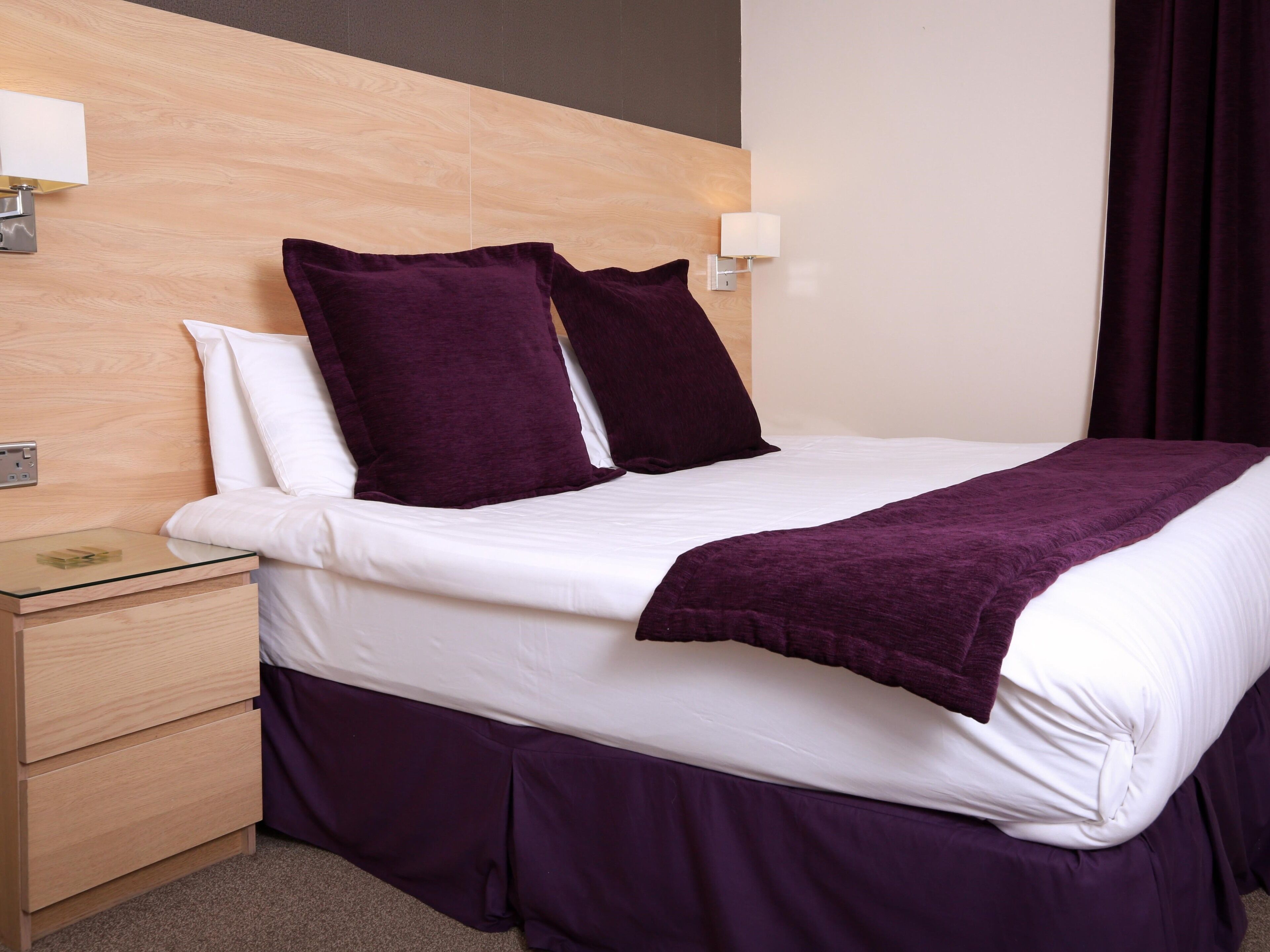 A neatly made bed with white linens and a purple blanket, a wooden nightstand with a glass top, and a wall-mounted light fixture.