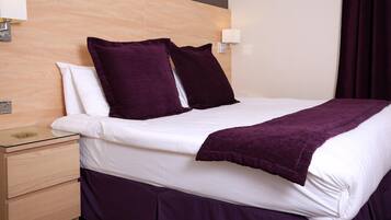 A neatly made bed with white linens and a purple blanket, a wooden nightstand with a glass top, and a wall-mounted light fixture.