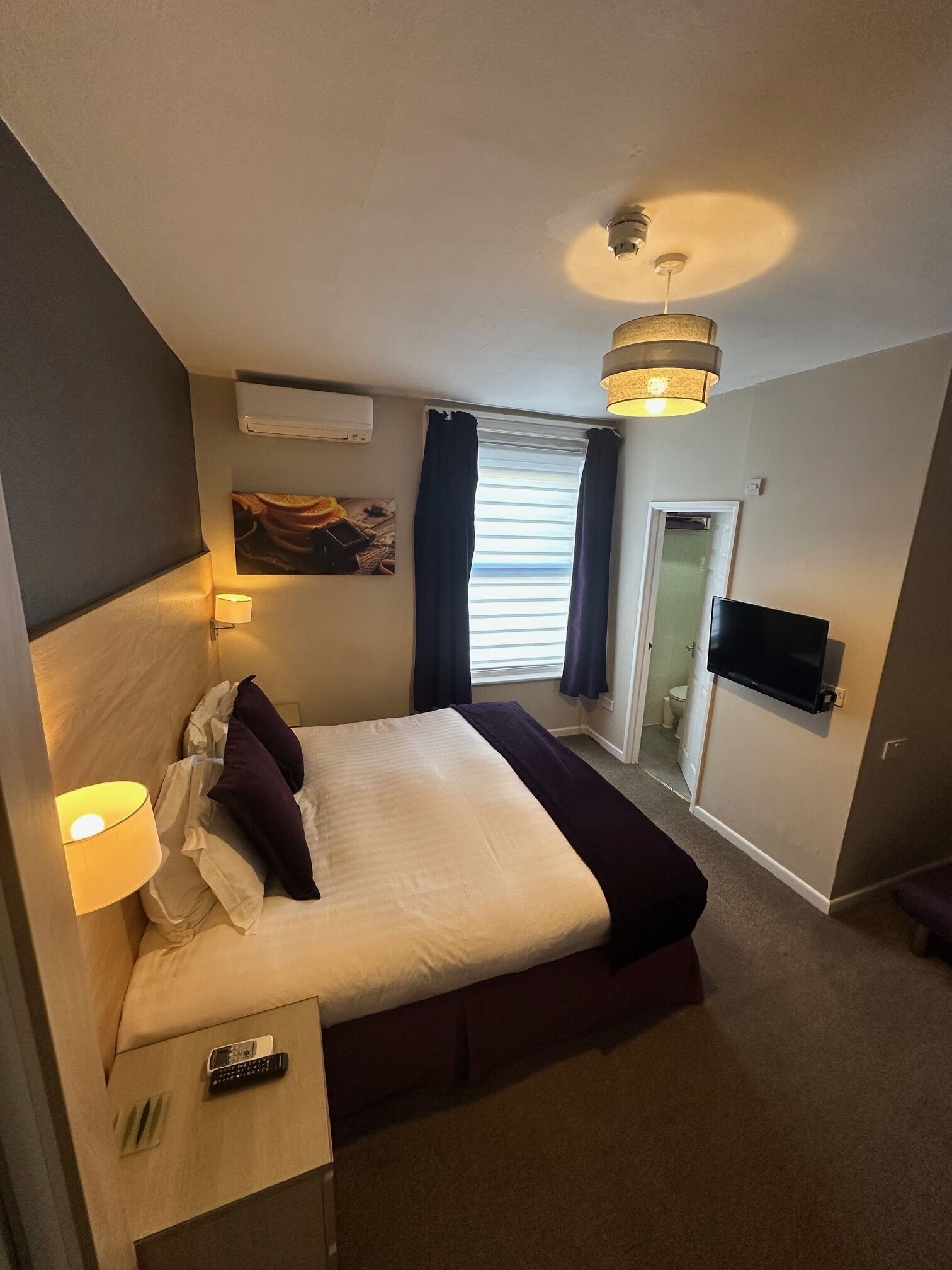 Une chambre d’hôtel comprenant un lit, une télévision, un luminaire au plafond, une fenêtre avec des rideaux et une porte donnant accès à une salle de bain.