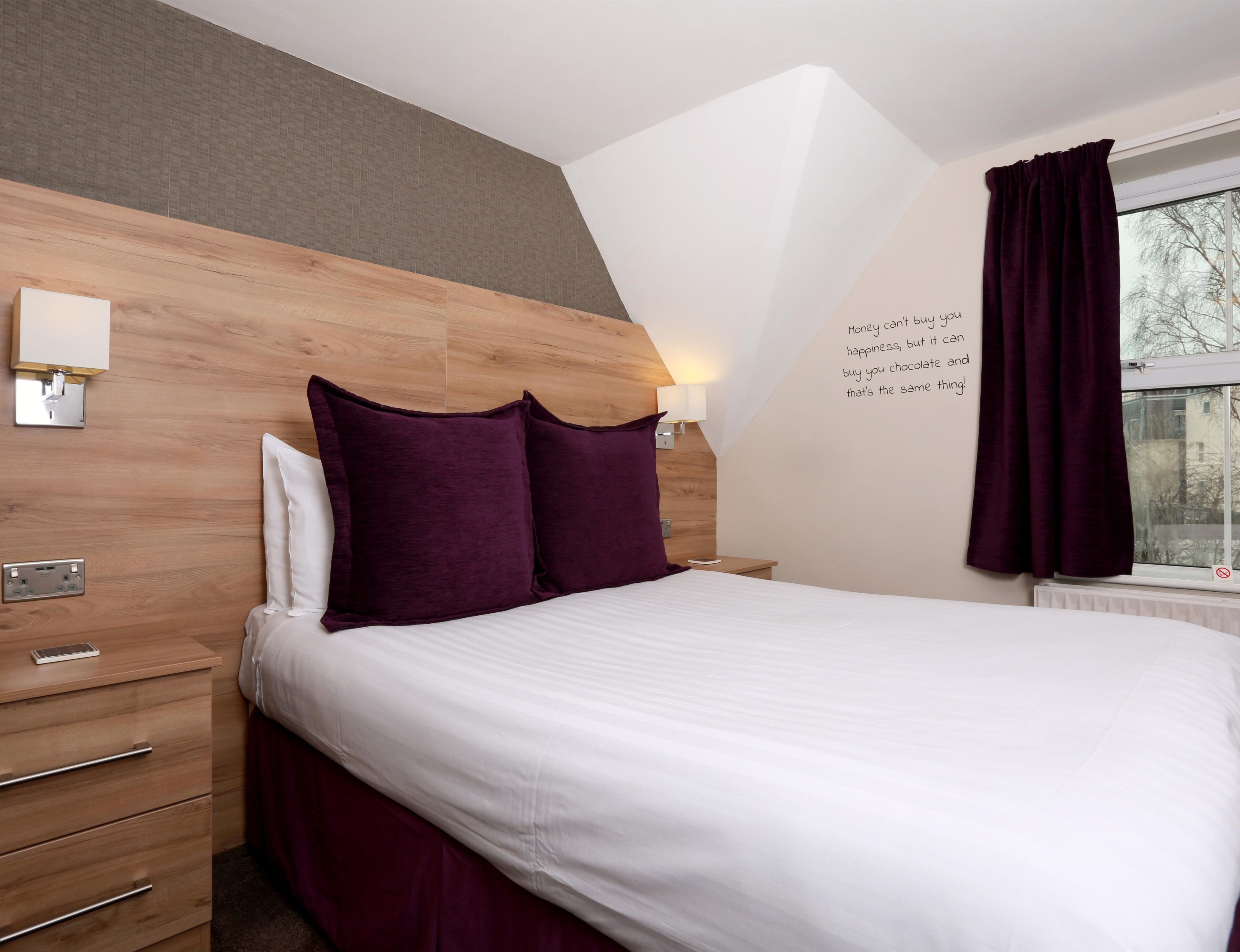 Une chambre moderne avec une tête de lit en bois, un lit blanc, des coussins violets, une table de chevet, une lampe et une fenêtre avec des rideaux.