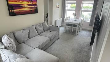 Uma sala de estar moderna com um sofá seccional cinza, uma mesa de jantar com cadeiras, uma TV de tela plana e uma grande janela com cortinas.