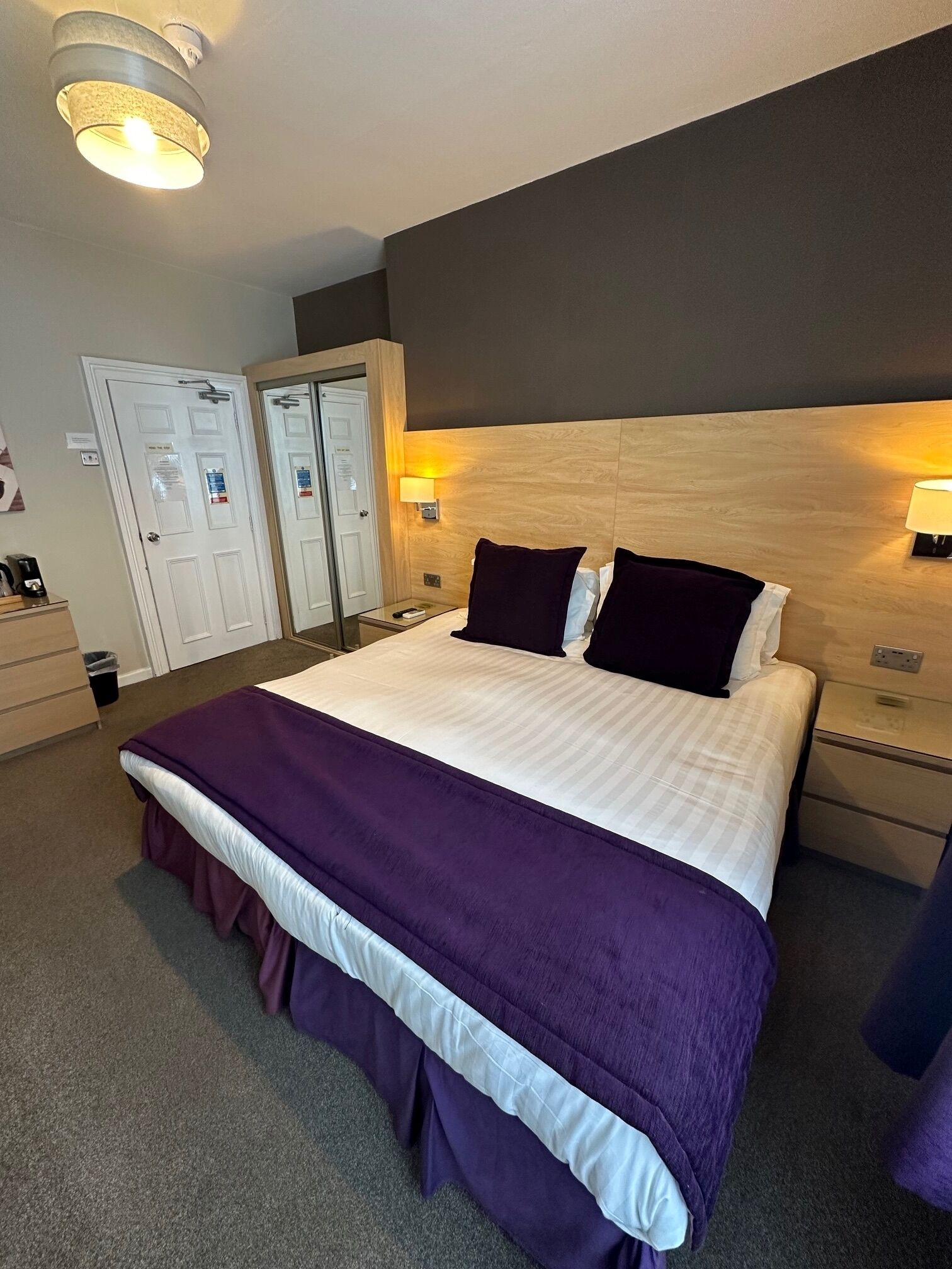 A hotel room with a large bed, two bedside tables, a nightstand, a wooden headboard, a ceiling light fixture, and a door with a mirror.