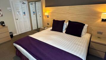 A hotel room with a large bed, two bedside tables, a nightstand, a wooden headboard, a ceiling light fixture, and a door with a mirror.