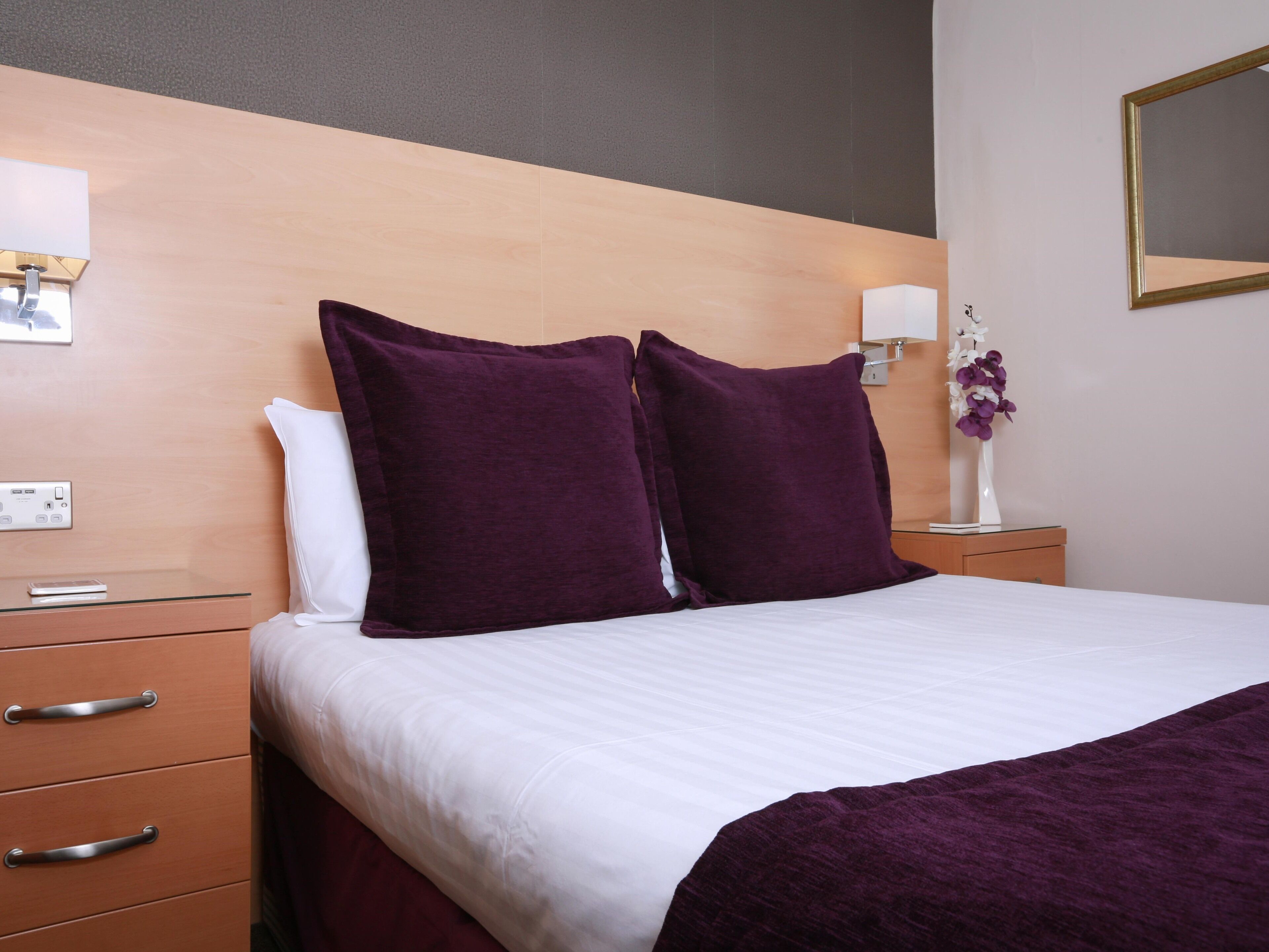 Een hotelkamer met een bed, twee kussens, een nachtkastje, een ladekast, een wandlamp en een vaas met bloemen.