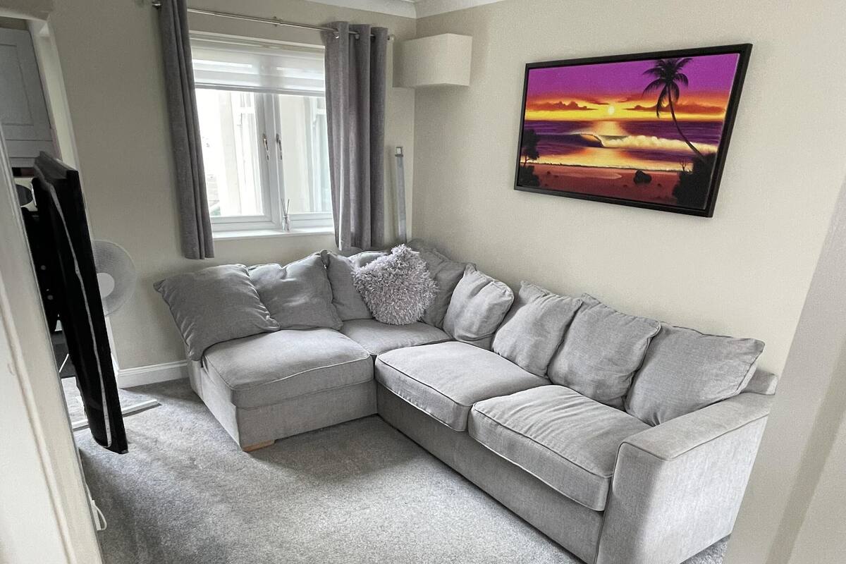Uma sala de estar com um sofá seccional cinza, um tapete cinza, uma pintura de um pôr do sol, uma janela com cortinas e uma porta com puxador de latão.