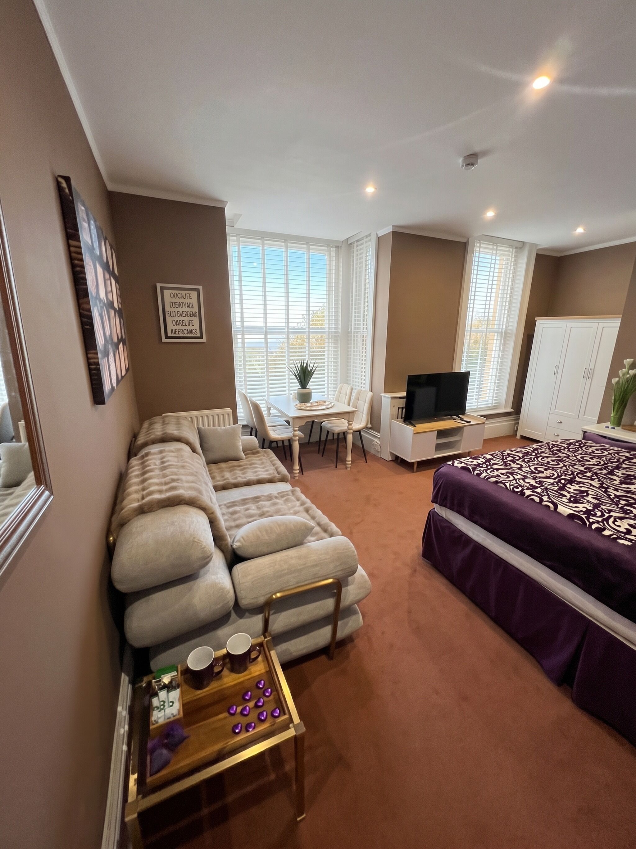 Una habitación con sofá modular, cama con colcha estampada, televisor, una mesita con adornos y ventana con persianas.
