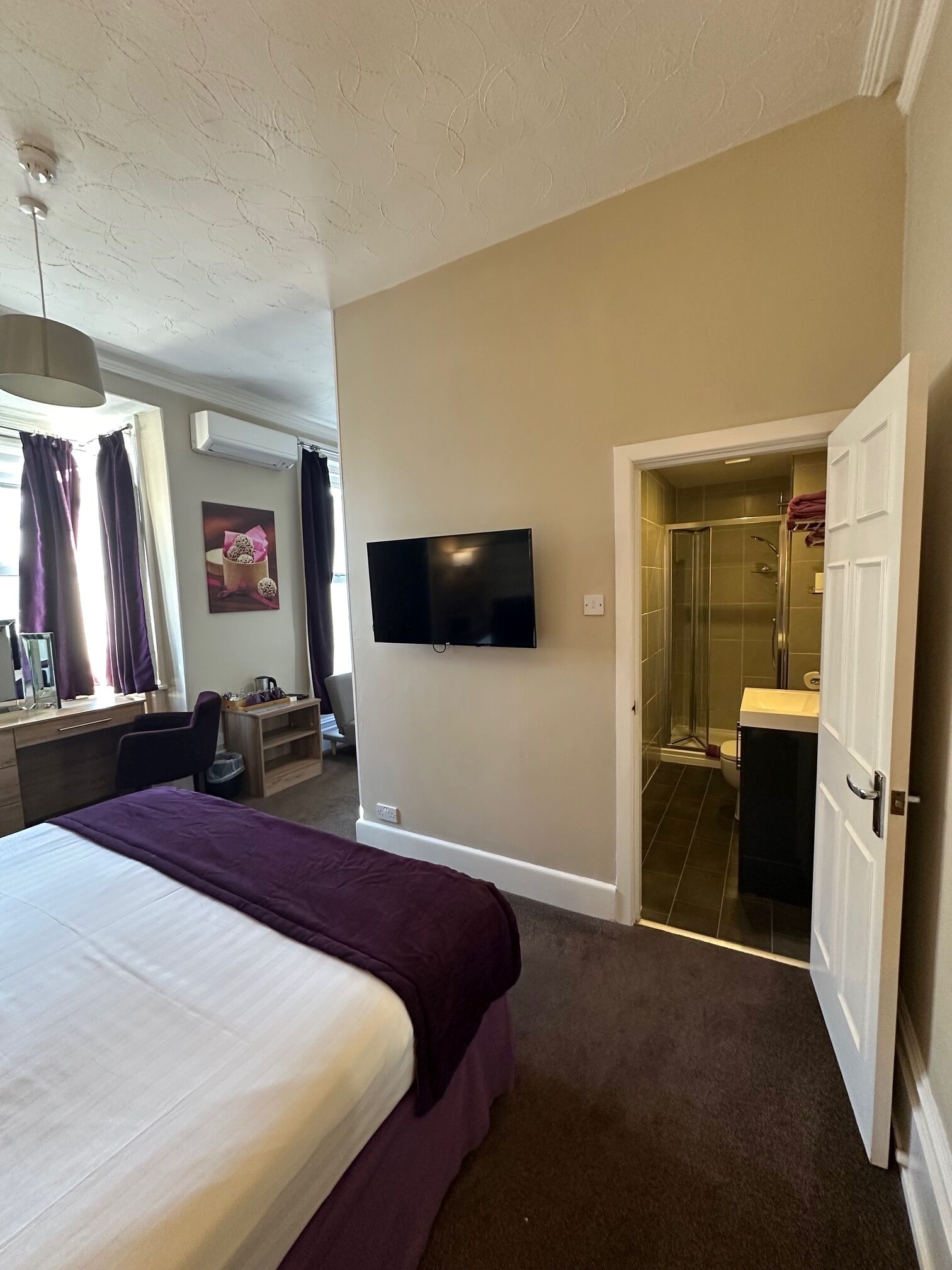A hotel room with a bed, a desk, a television, an air conditioning unit, a shower, and a bathroom.