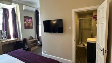 A hotel room with a bed, a desk, a television, an air conditioning unit, a shower, and a bathroom.