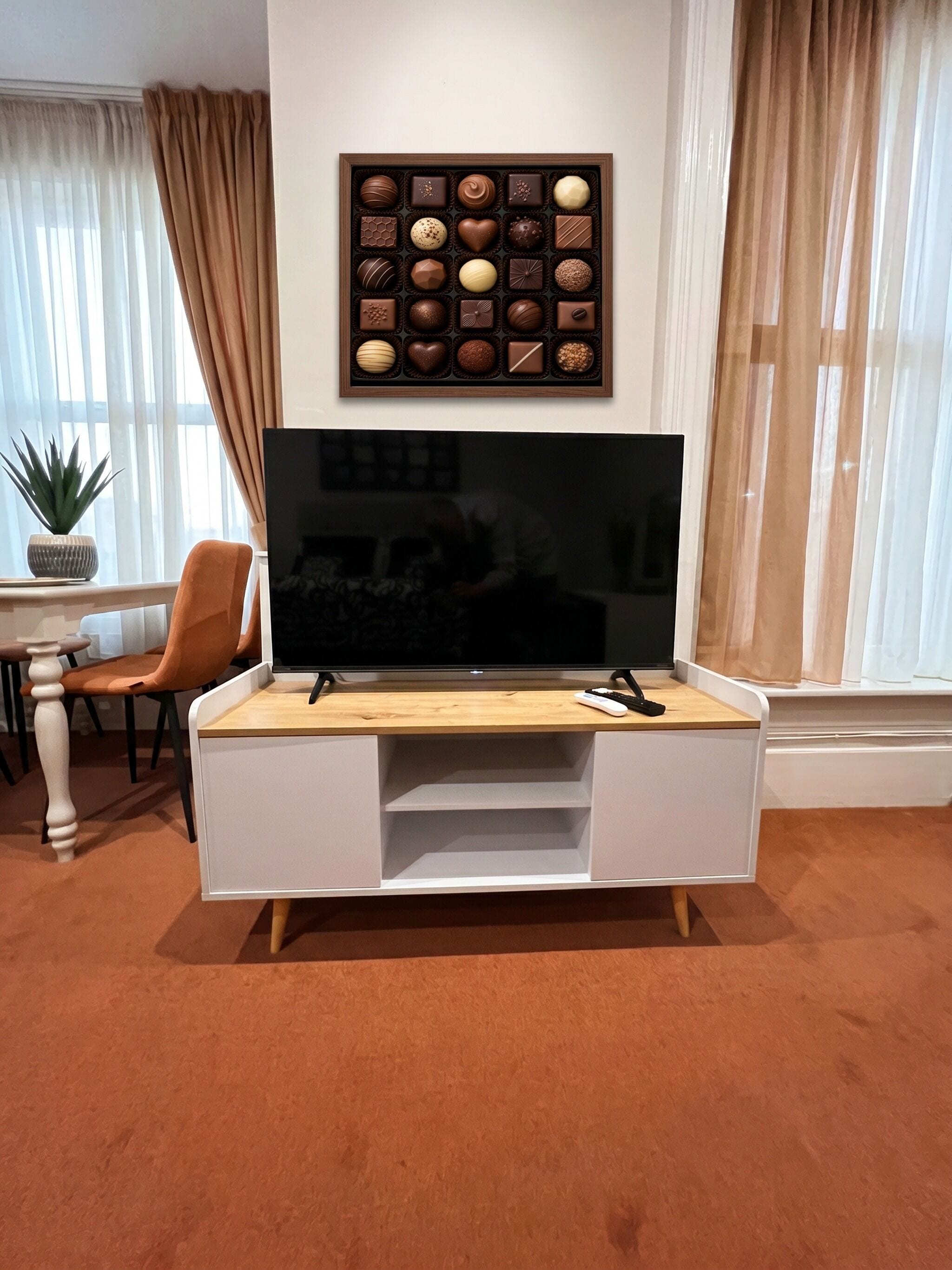 A living room with a flat-screen TV on a white entertainment unit, a potted plant, and a framed chocolate box on the wall.