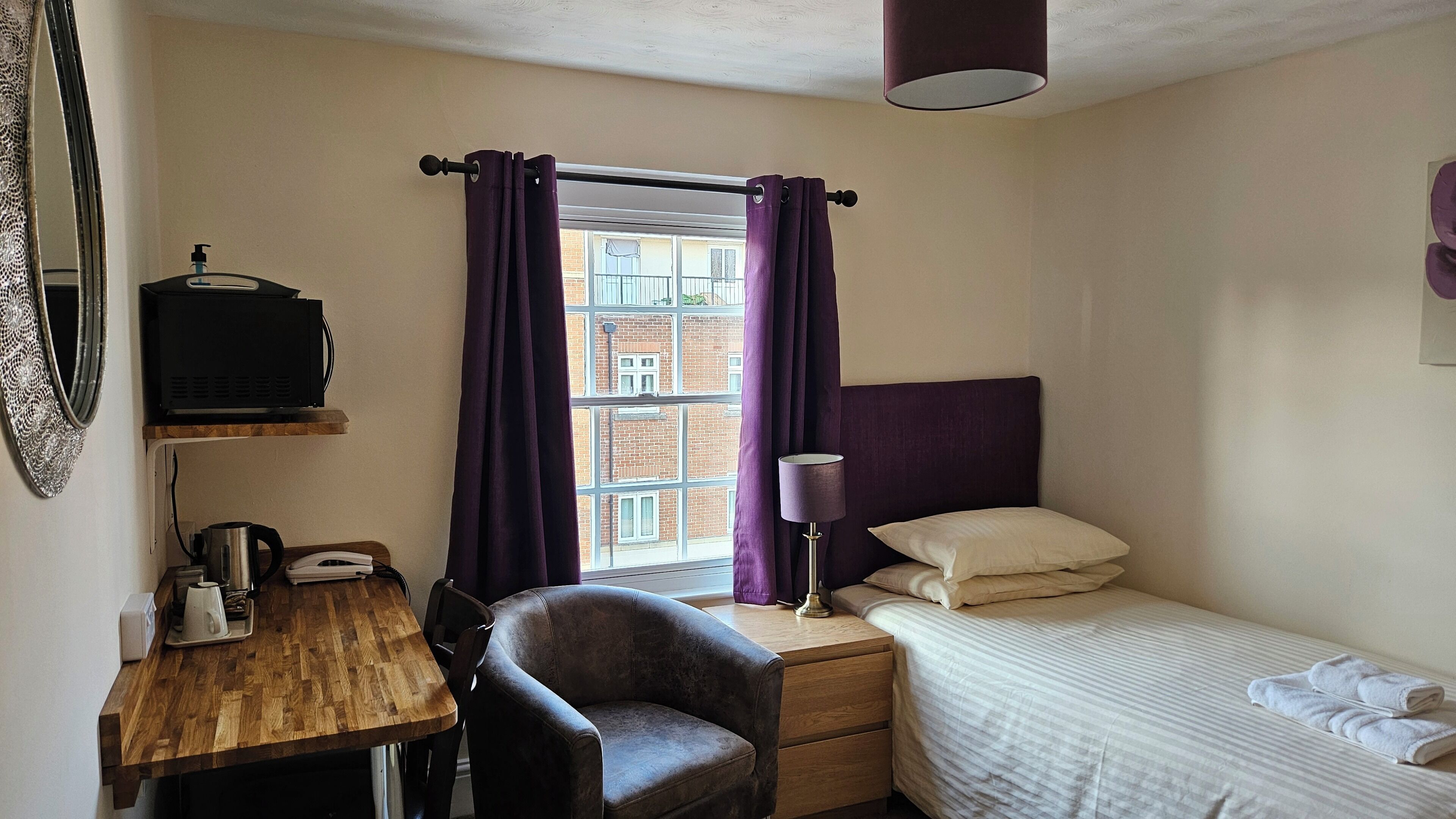 A small, neatly arranged bedroom with a wooden desk, a chair, a bed, and a window with purple curtains.