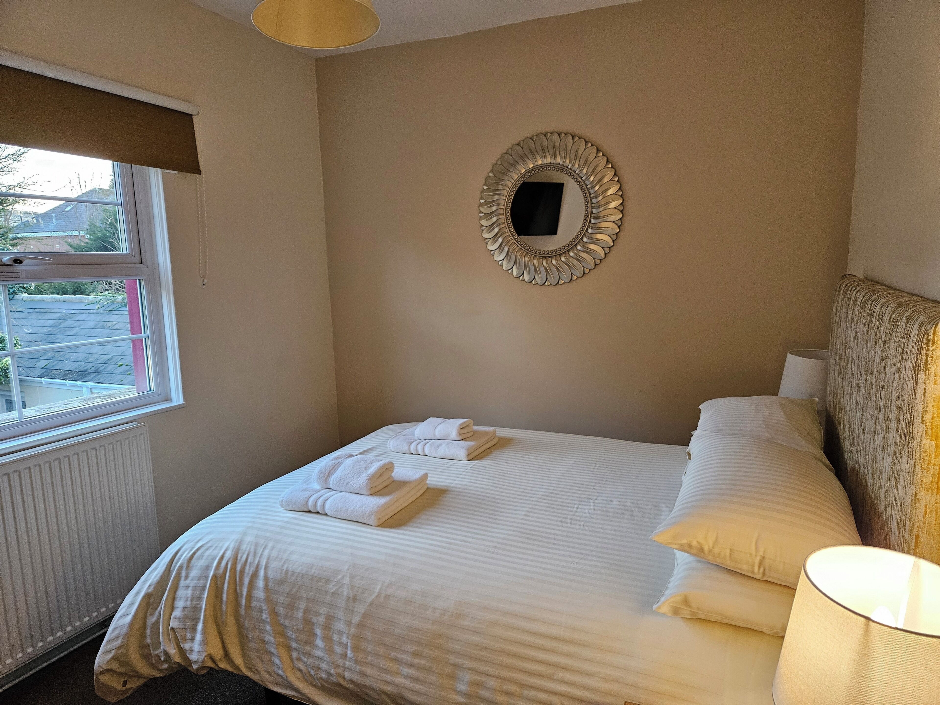 A hotel room with a bed, a window, a radiator, and a wall-mounted mirror.