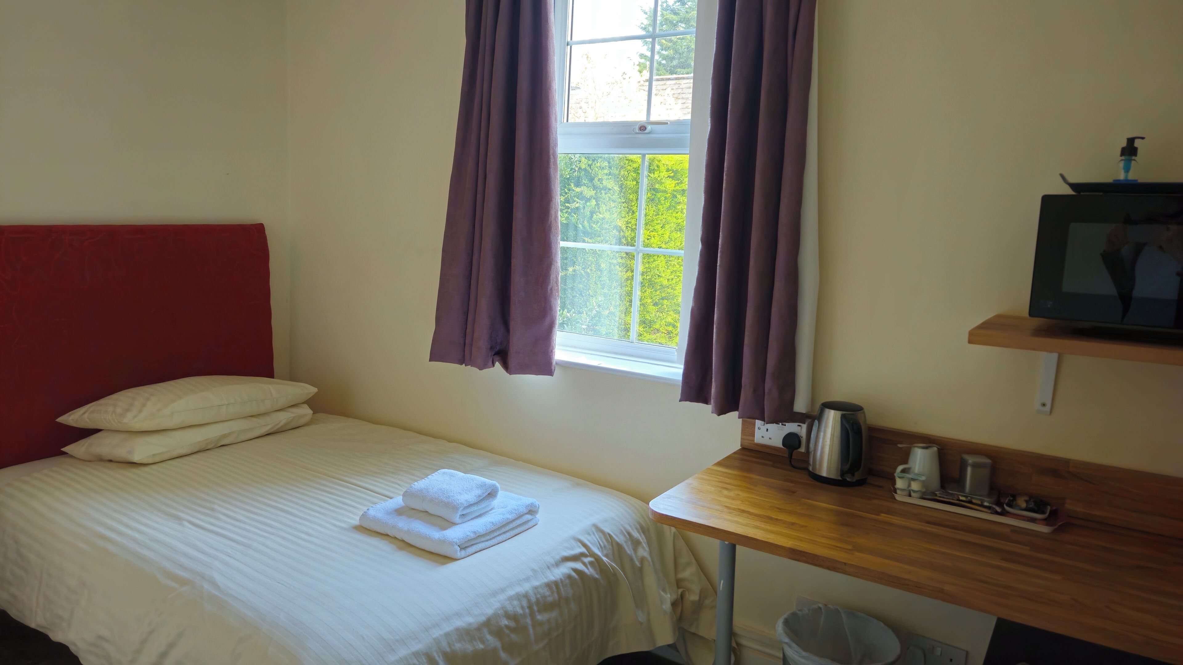 A single bed with white bedding and a red headboard, a wooden desk with a television, a kettle, and a cup, and a window with purple curtains.