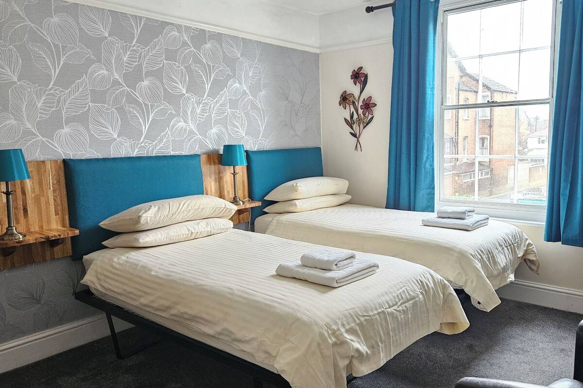 A hotel room with two beds, a window with blue curtains, a wall-mounted decorative piece, and a wooden headboard.
