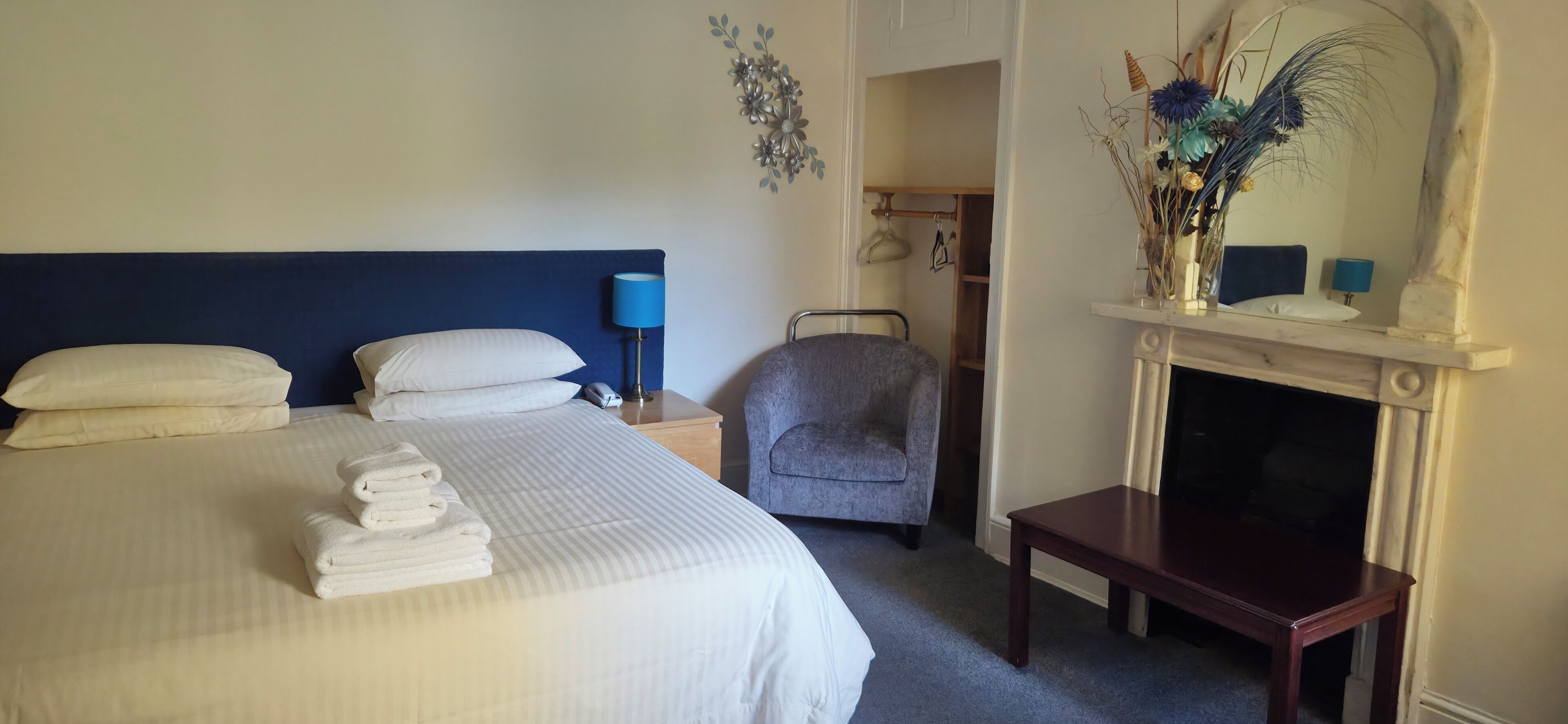 A hotel room with a large bed, a chair, a small table, a nightstand, a lamp, and a fireplace.