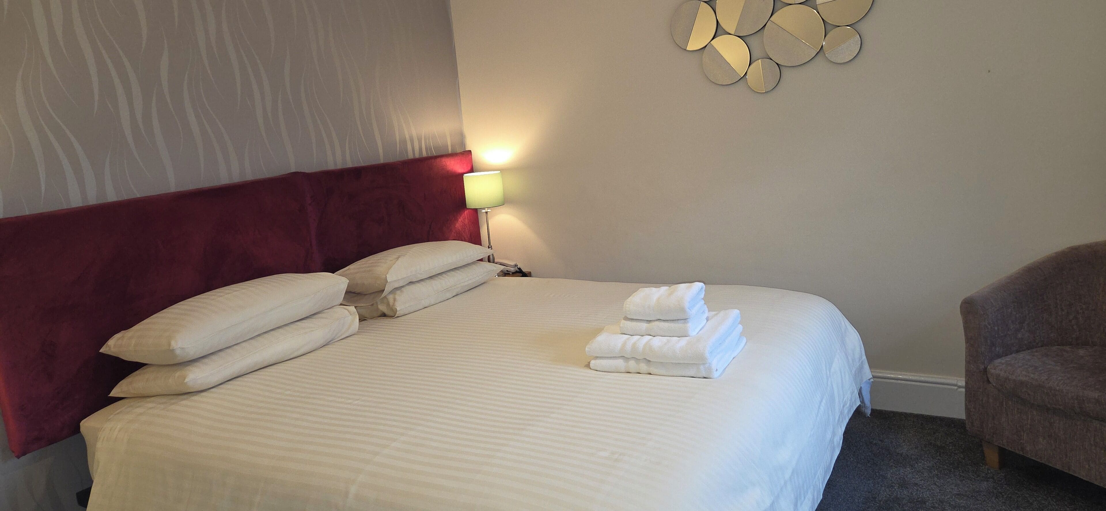 A neatly made bed with white linens and pillows, a red headboard, a wall-mounted light, and a chair to the right.