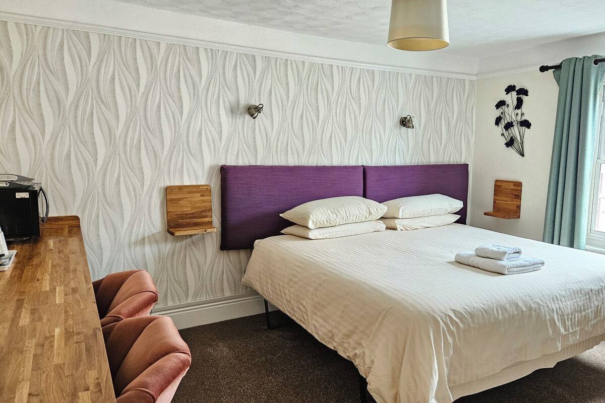 A neatly arranged bedroom with a bed, a wooden side table, a chair, a wall-mounted lamp, and a window with curtains.