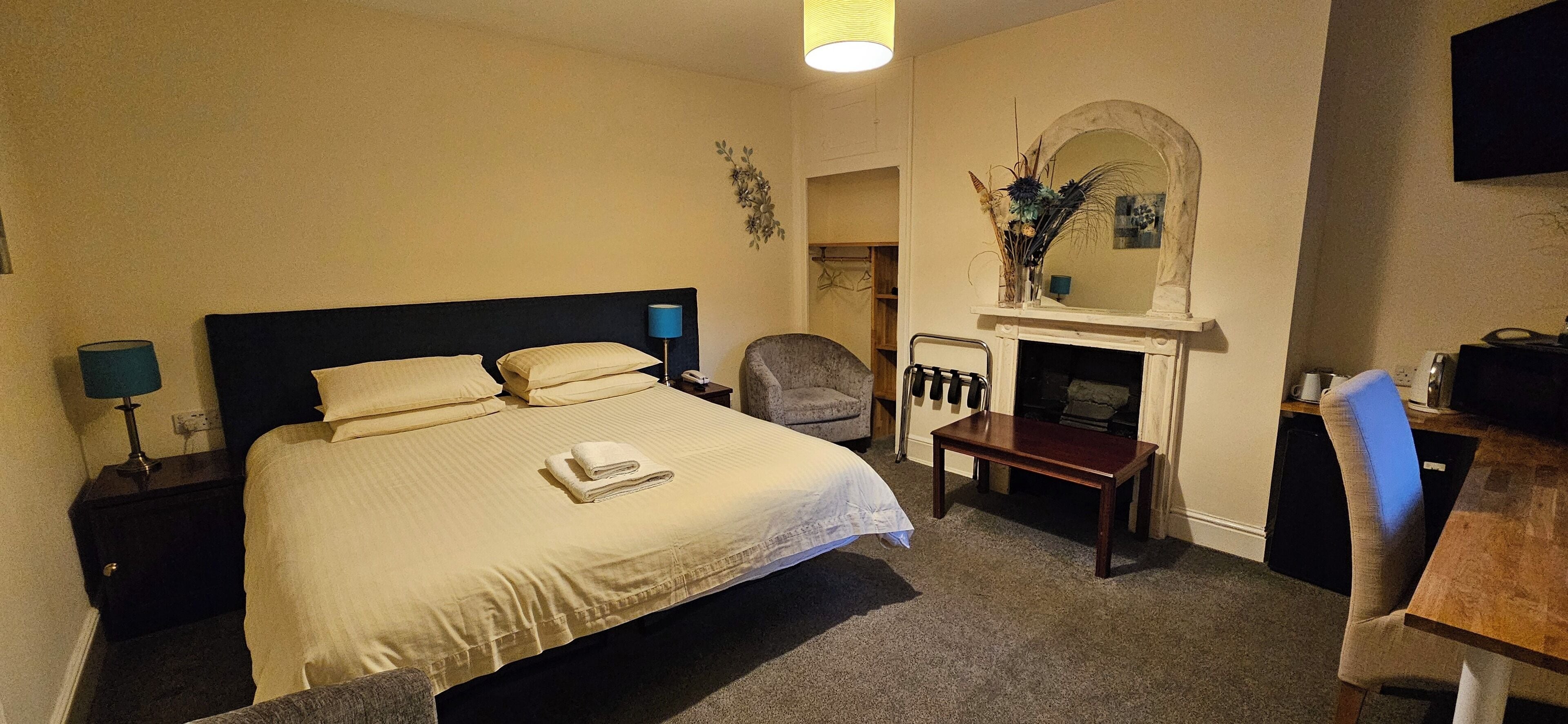 A hotel room with a large bed, a small table, a chair, a desk, a fireplace, and a TV.