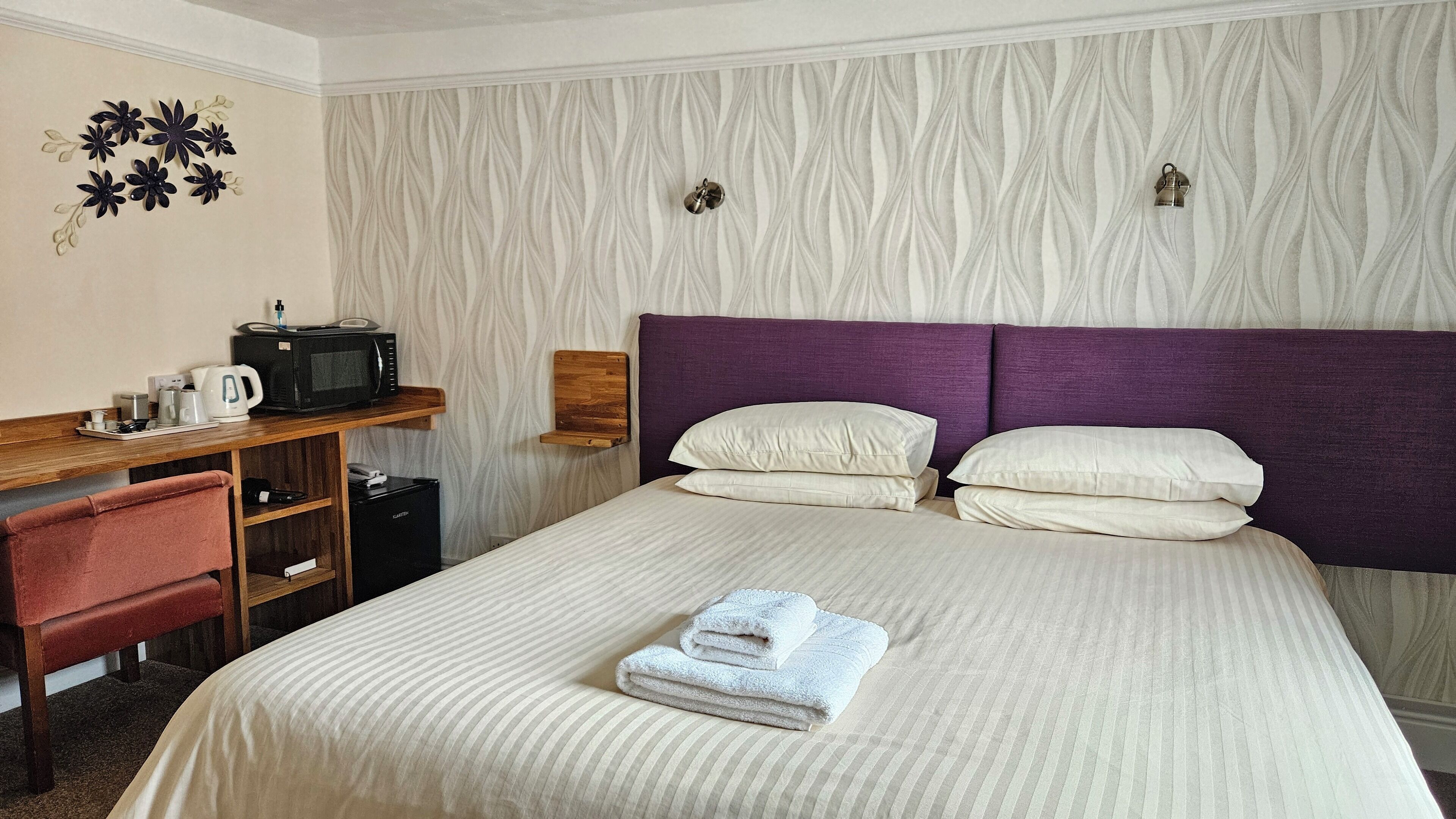 Ett hotellrum med en säng, ett skrivbord med en mikrovågsugn, en stol och en hylla som är monterad på väggen.