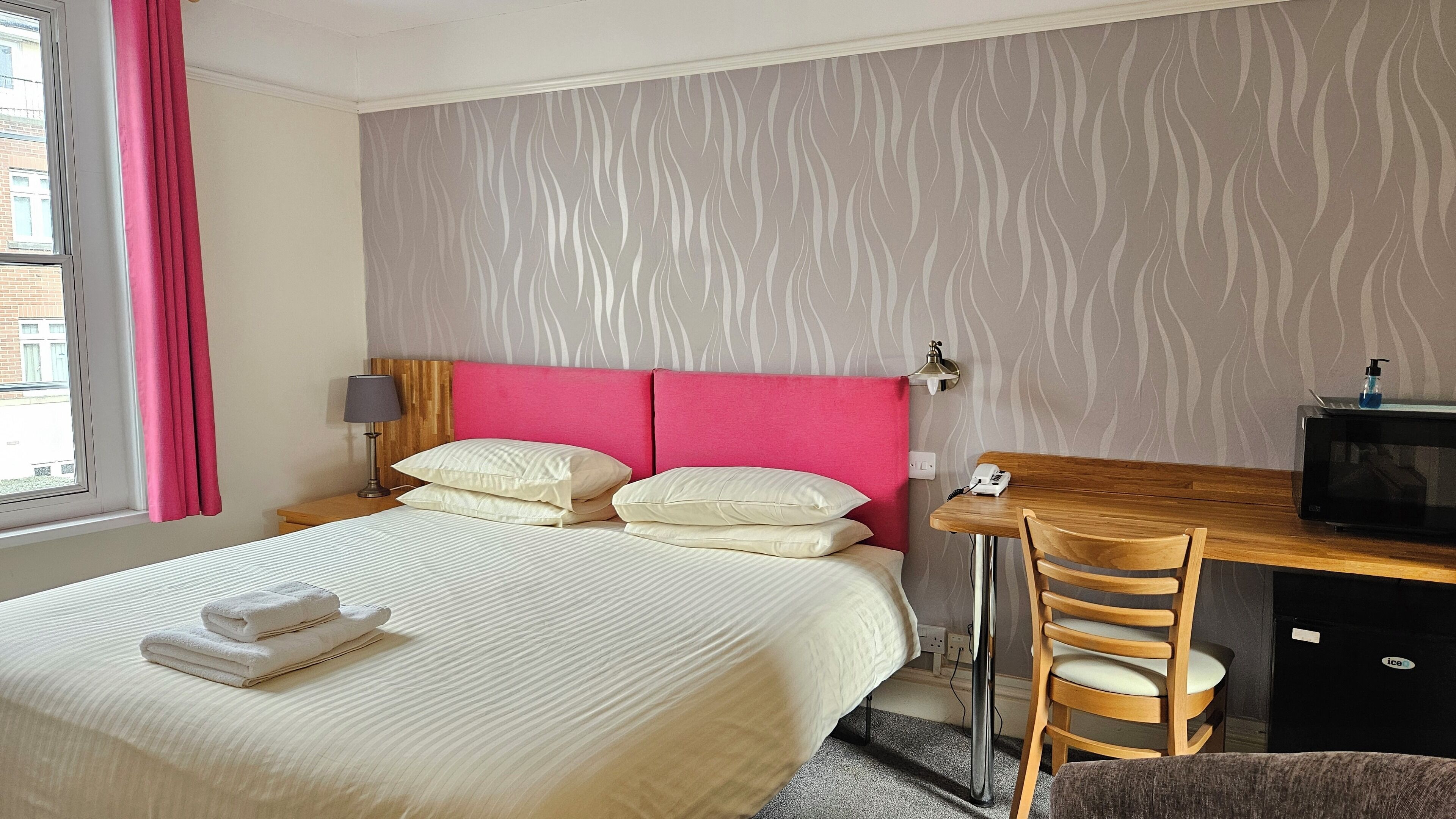 Habitación de hotel con cama, almohadas, escritorio con silla, lámpara, teléfono, microondas y ventana con cortinas.
