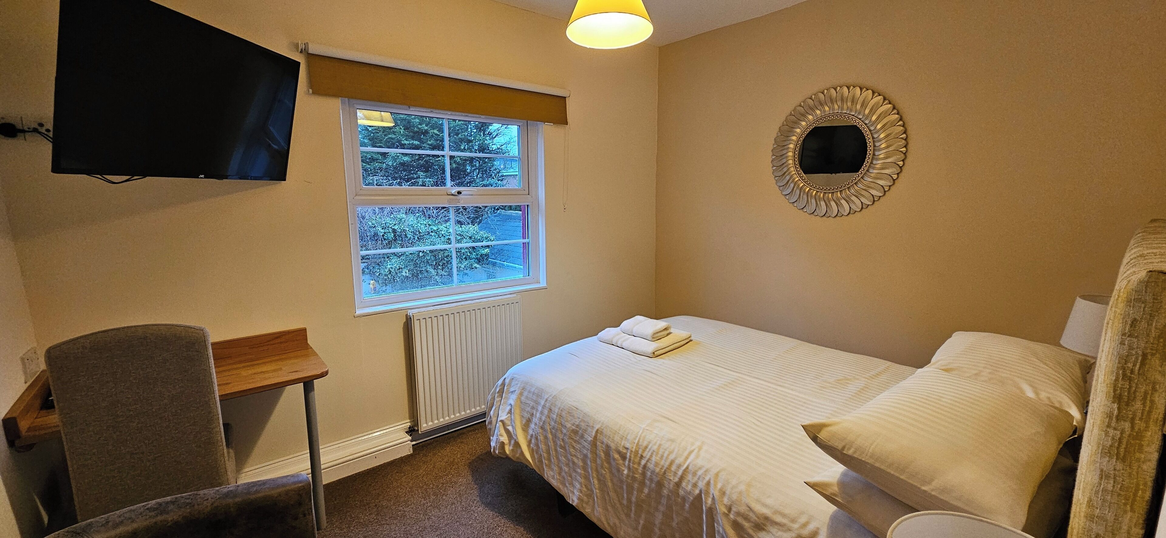 A bedroom with a bed, a chair, a desk, a television, a window, and a radiator.