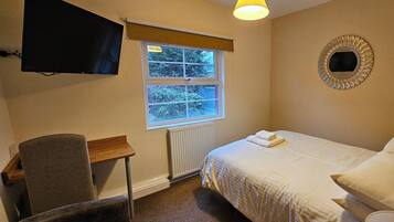 A bedroom with a bed, a chair, a desk, a television, a window, and a radiator.