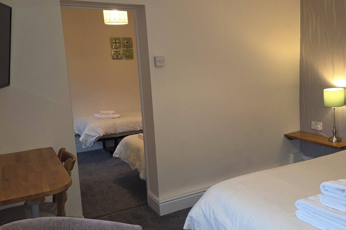 A hotel room with a bed, a bedside table with a lamp, a chair, a desk, and a small table with a kettle.