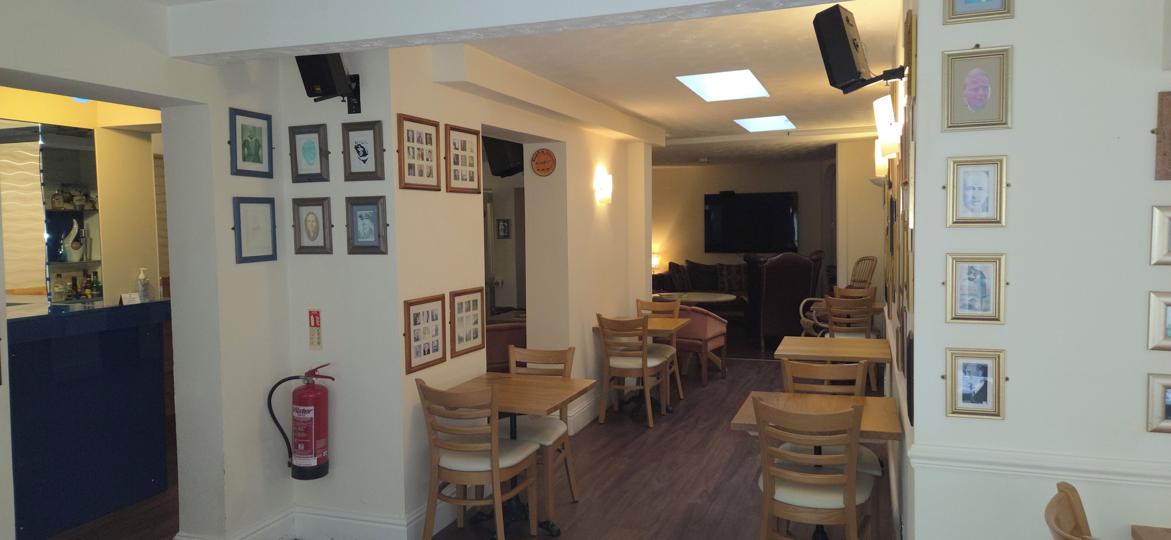 A dining area with wooden tables and chairs, framed pictures on the walls, and a fire extinguisher.