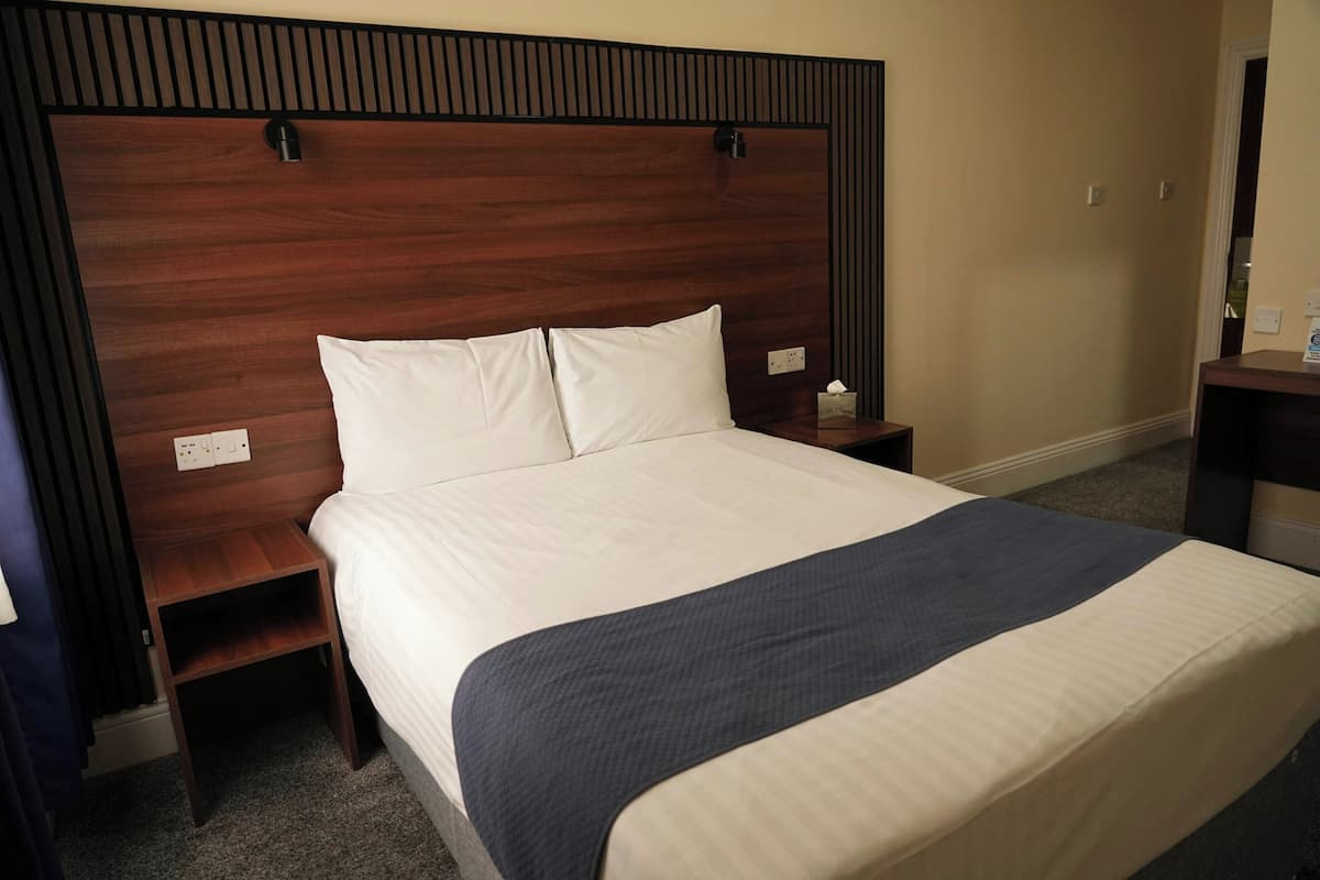 A hotel room with a wooden headboard, a large bed with white bedding, a bedside table with a lamp, and a nightstand.