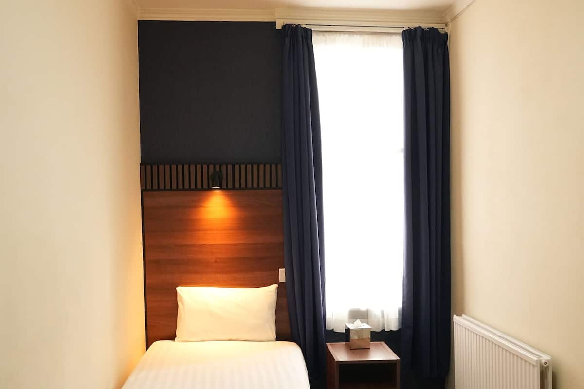A small, neatly arranged hotel room with a single bed, a bedside table, and a window with curtains.