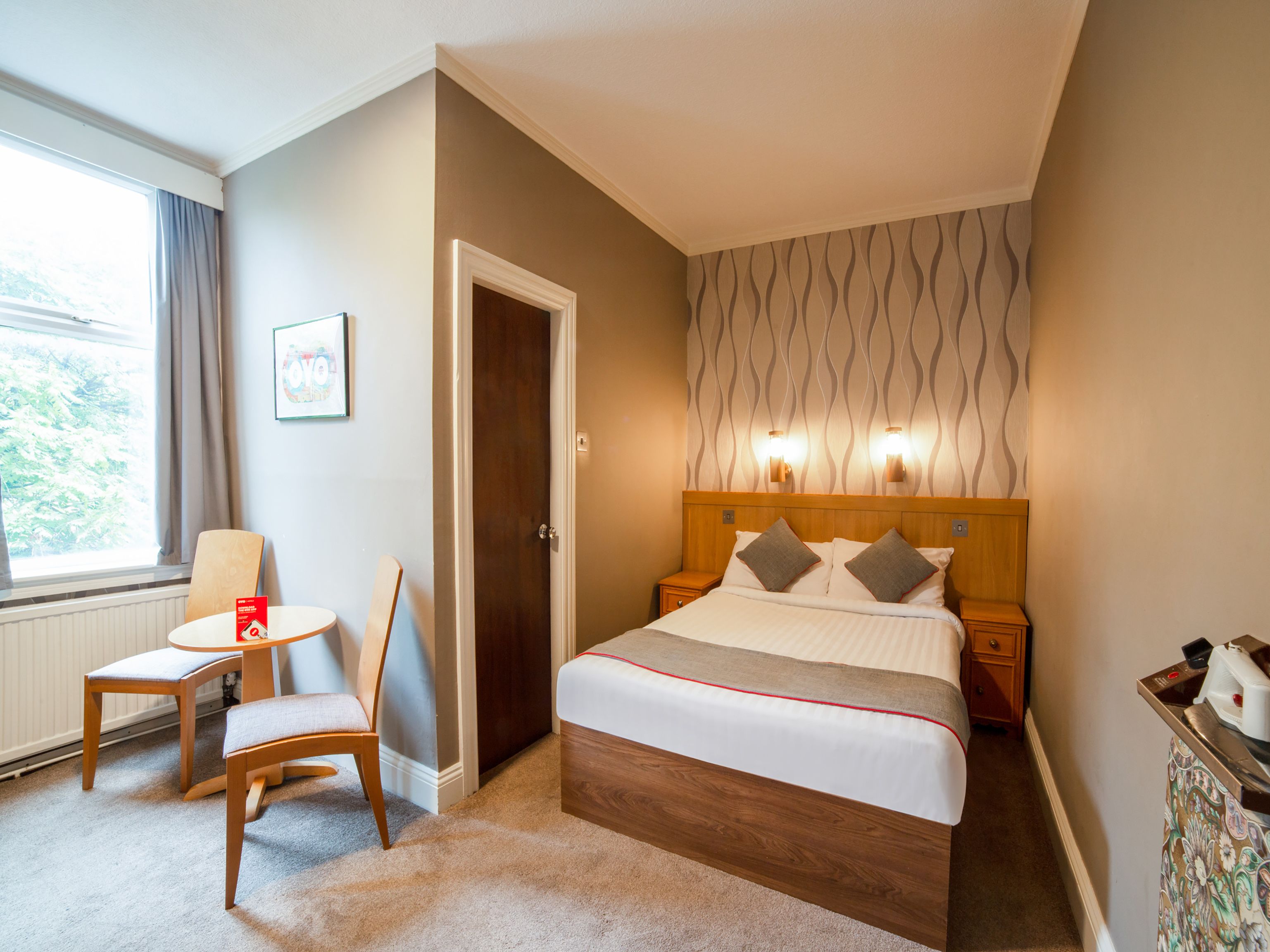 A hotel room with a bed, bedside tables, a chair, a small table, and a window with curtains.