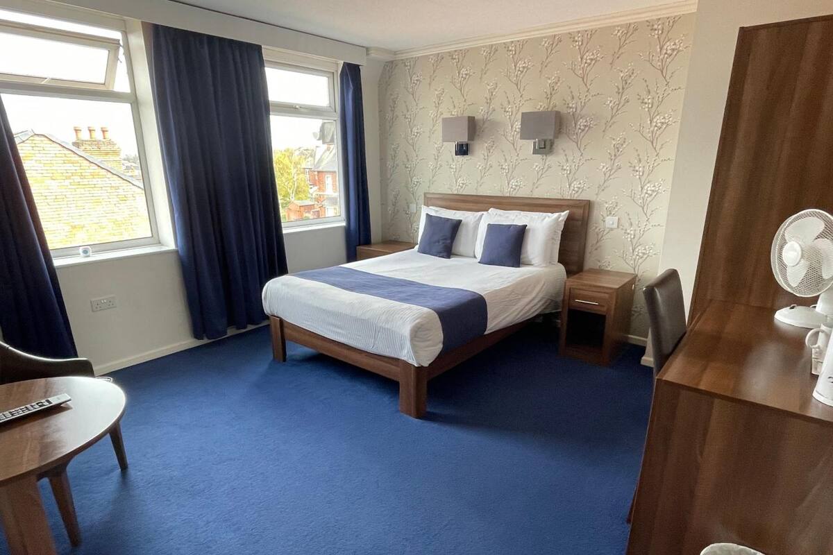 A hotel room with a bed, bedside tables, a desk, a chair, a window with curtains, and a wall-mounted fan.
