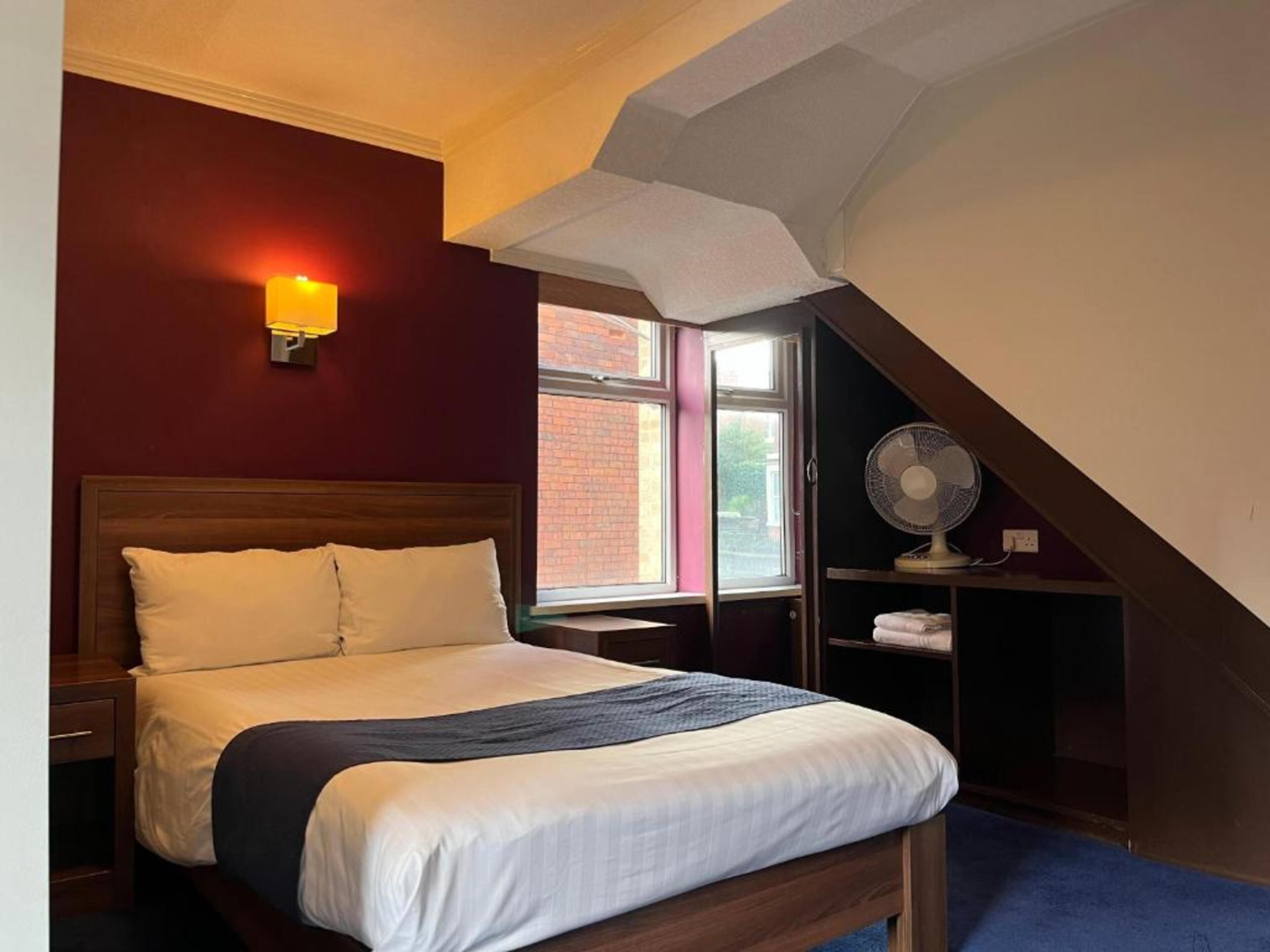 A bedroom with a bed, a nightstand, a wall-mounted lamp, a window, and a fan.