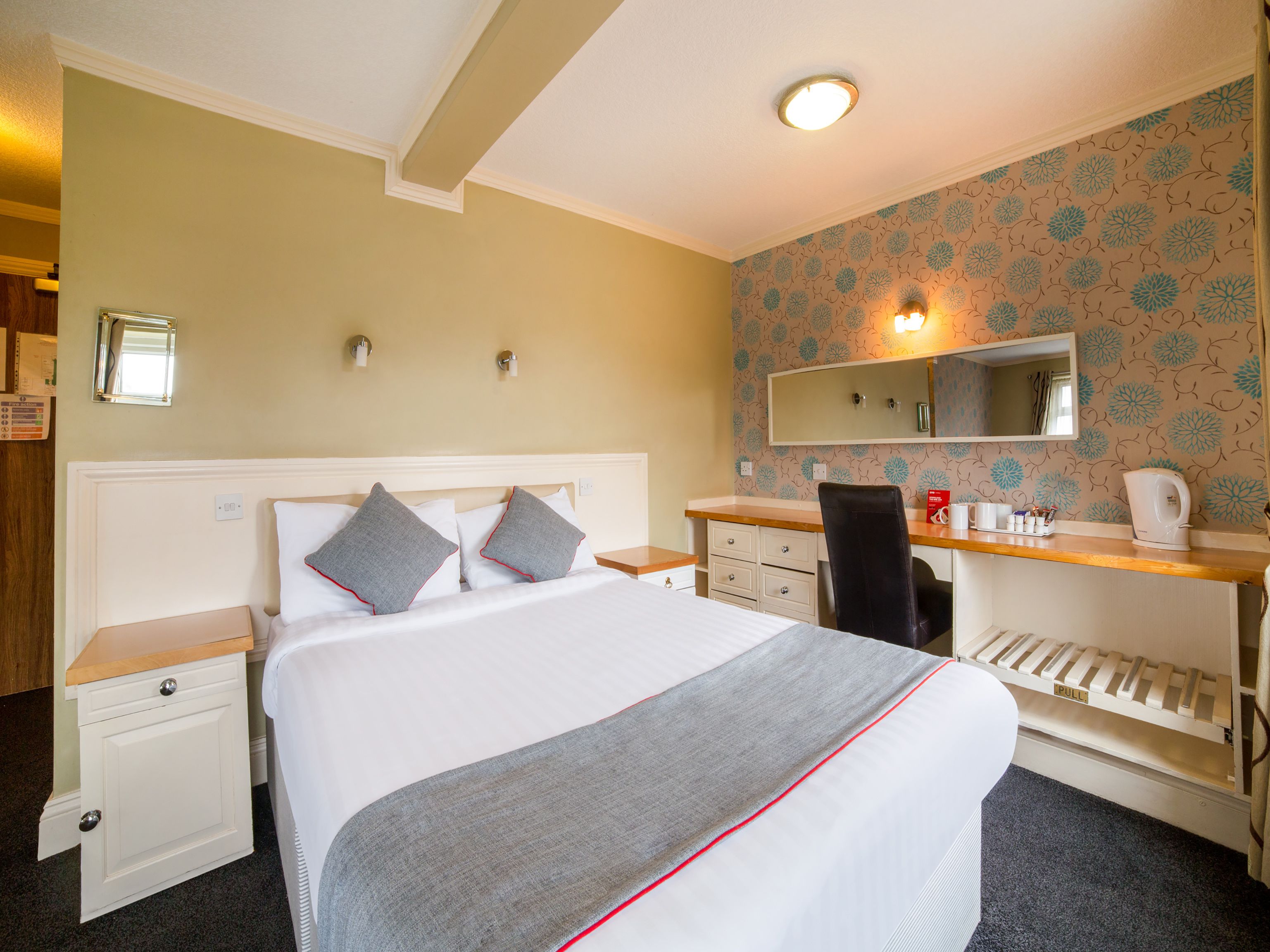 A hotel room with a bed, a desk, a chair, a mirror, and a small kitchenette.