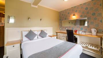 A hotel room with a bed, a desk, a chair, a mirror, and a small kitchenette.