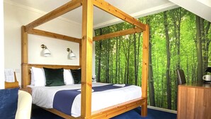 A four-poster bed with a blue carpet, a desk with a coffee maker, and a forest wallpaper background. - The Continental Hotel, Derby (Derby)