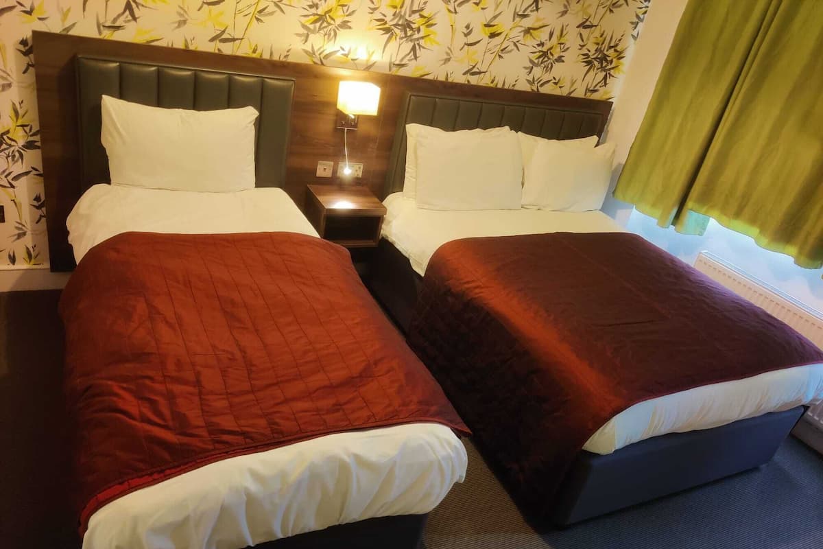 A hotel room with two beds, each with a dark headboard and white bedding. There is a nightstand with a lamp between the beds.