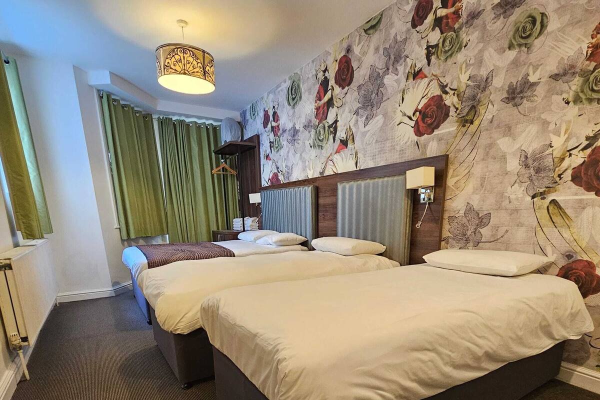 A hotel room with two beds, a large floral wallpaper, a hanging light fixture, and a green curtain.