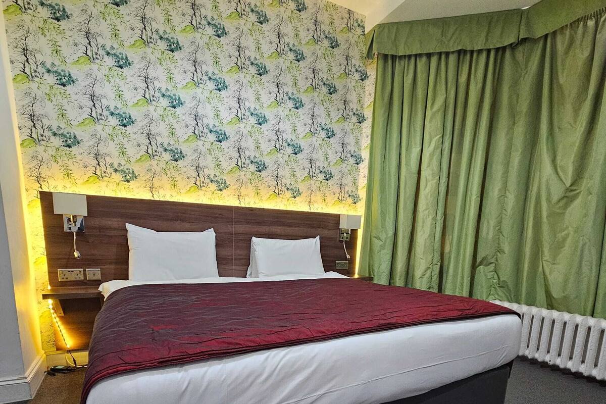 A hotel room with a large bed, a headboard with a floral wallpaper design, a green curtain, and a nightstand with a lamp.