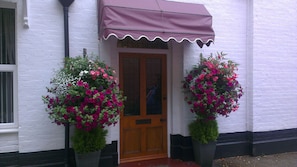 A white building with a wooden door and a brown awning. There are two large potted plants with pink flowers on either side of the door. - The Rosscourt - Adults only (Bournemouth)