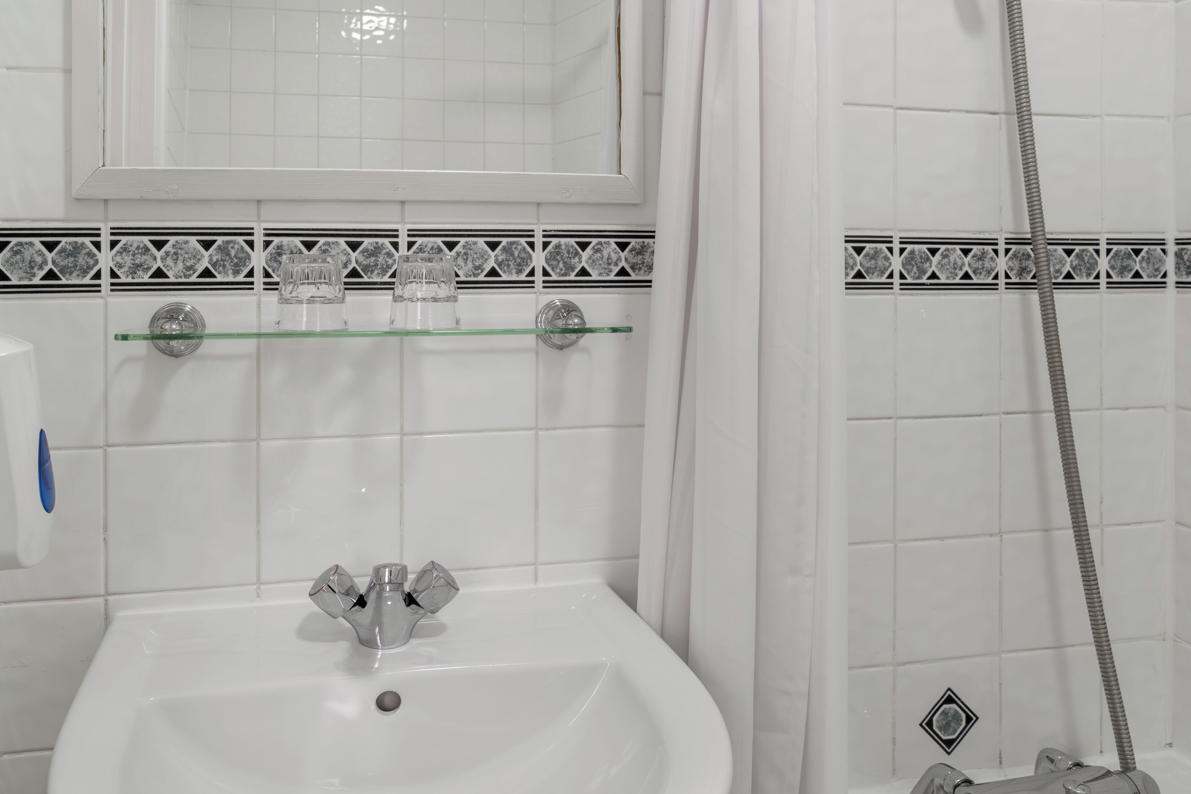 Une salle de bain avec un lavabo blanc, une étagère en verre au-dessus, une douche avec un rideau et un mur carrelé orné d’une bordure décorative.