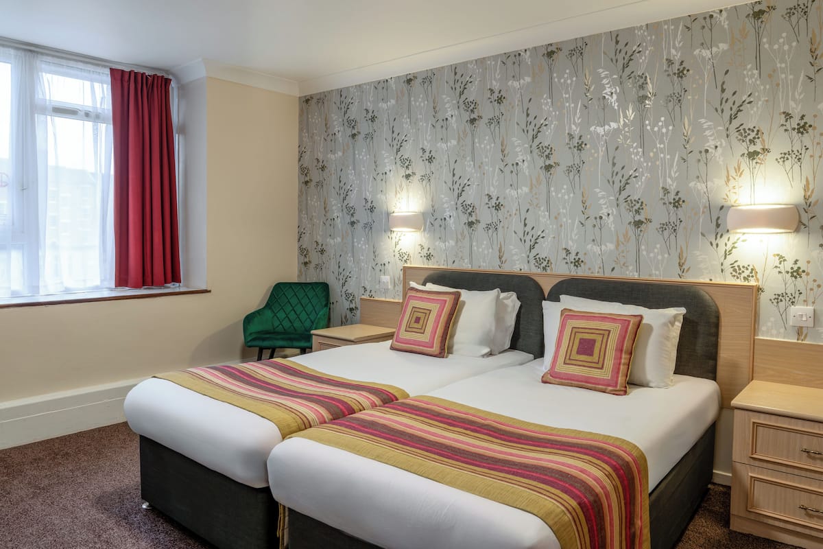 A double bed with two pillows, a bedside table, a nightstand, a green chair, and a window with red curtains.
