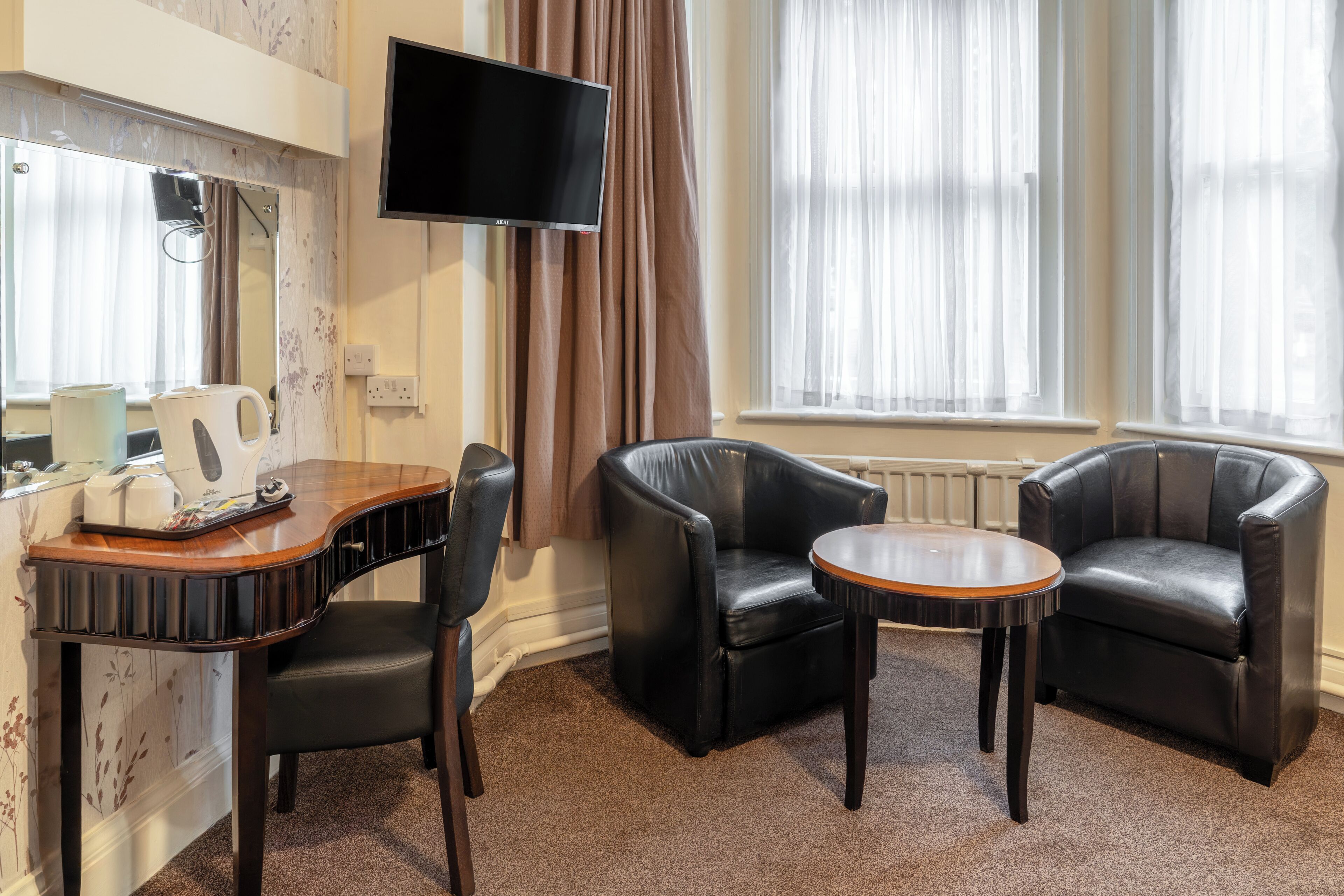 A room with a desk, two leather chairs, a round table, a flat-screen TV, and a window with curtains.