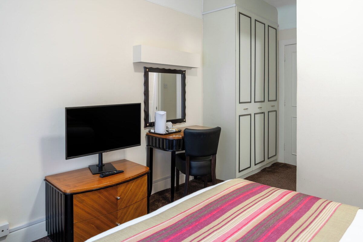 A hotel room with a bed, a television on a wooden dresser, a desk with a chair, a mirror, and a wardrobe.