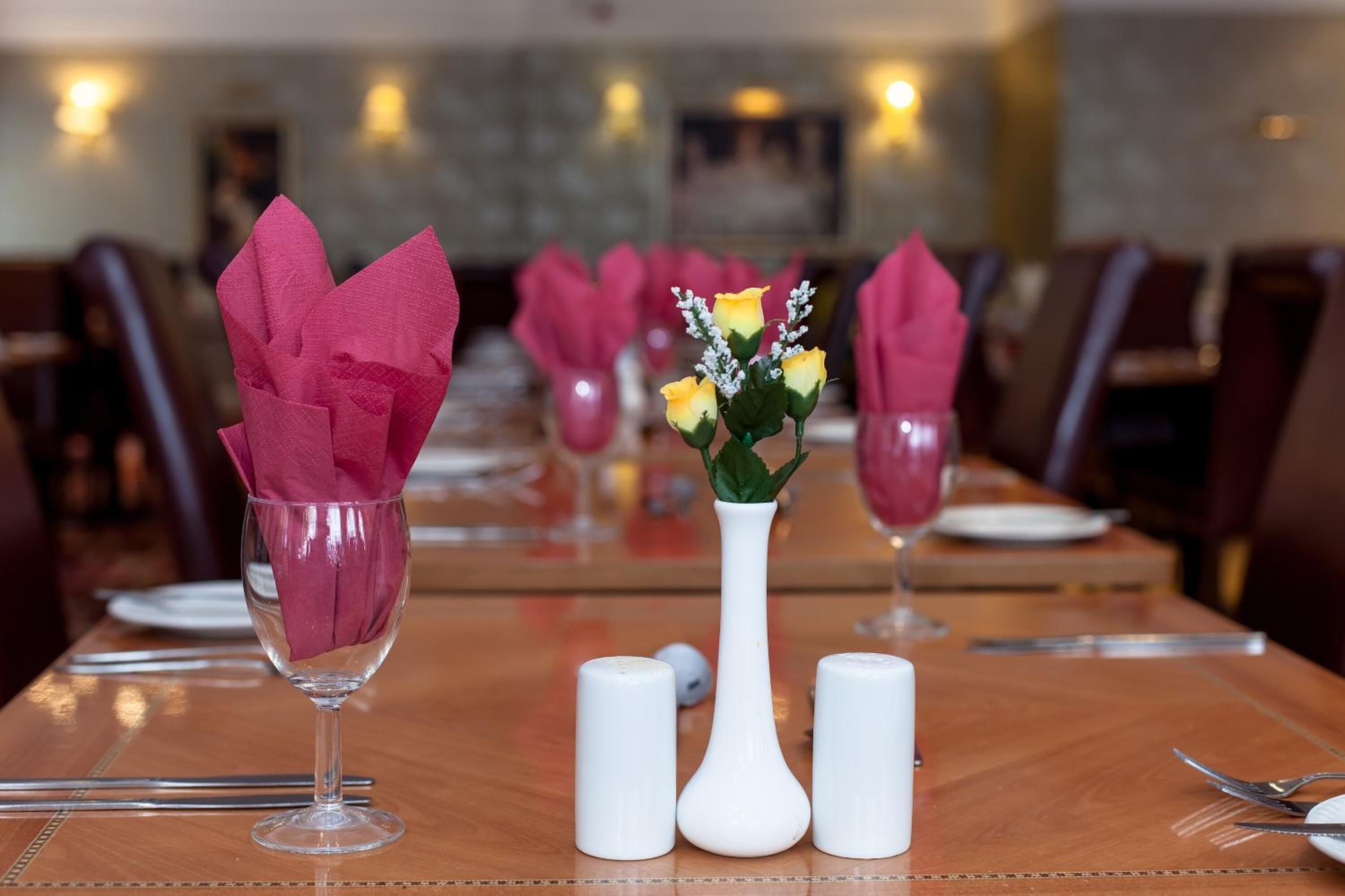 Een eettafel gedekt met roze servetten, een witte vaas met gele bloemen en zout- en peperstrooiers.