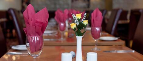 Een eettafel gedekt met roze servetten, een witte vaas met gele bloemen en zout- en peperstrooiers.