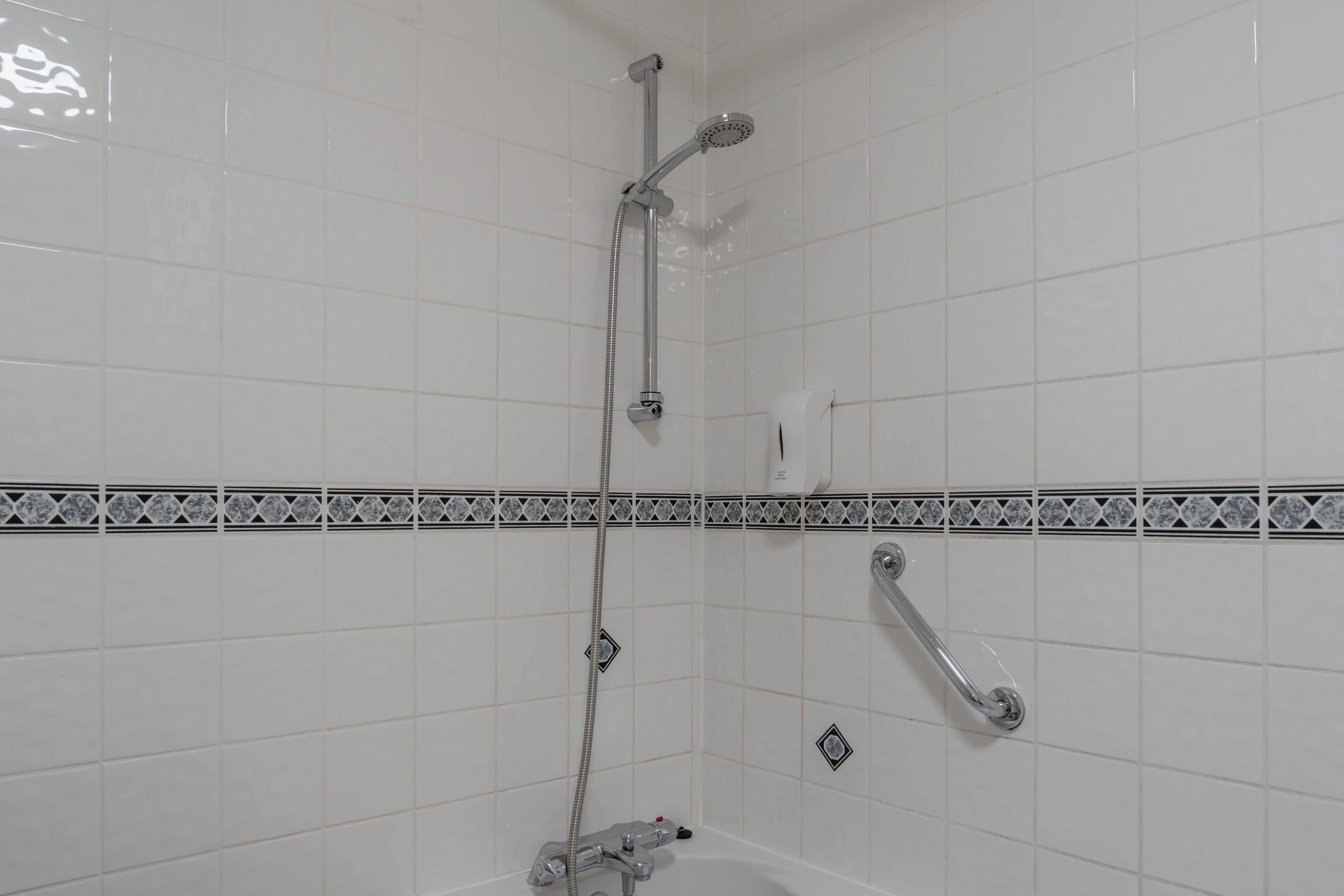 A bathroom with white tiled walls, a shower with a handheld showerhead, and a handheld faucet.