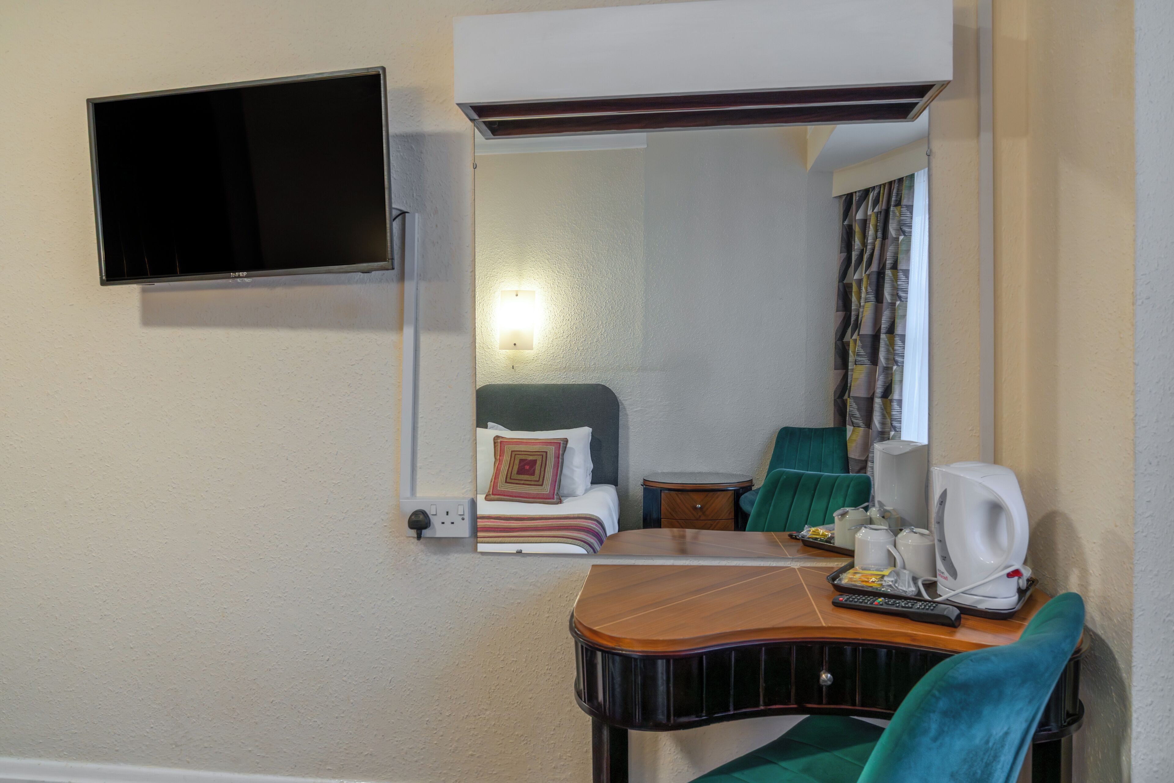 Een hotelkamer met een bed, een bureau met stoel, een aan de muur bevestigde televisie en een tafeltje met waterkoker en kopjes.
